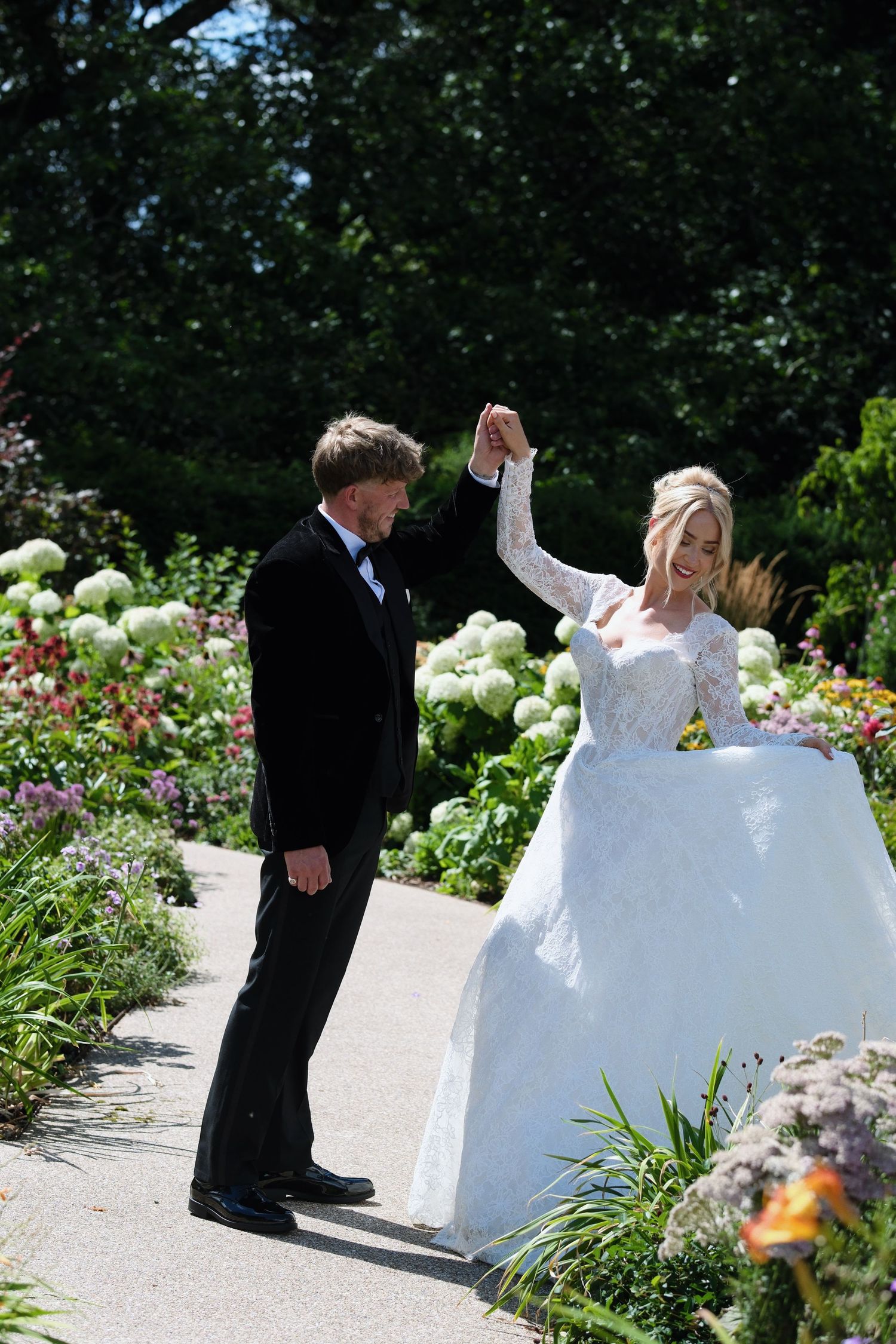 Bride and groom dancing joyfully in a lush garden, bride in white lace gown, groom in black tuxedo.
