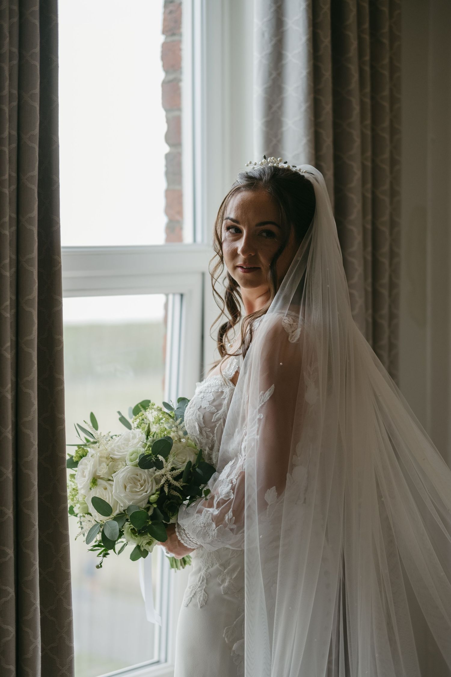 Bride in lace gown and veil holds white rose bouquet by window in elegant bridal portrait.