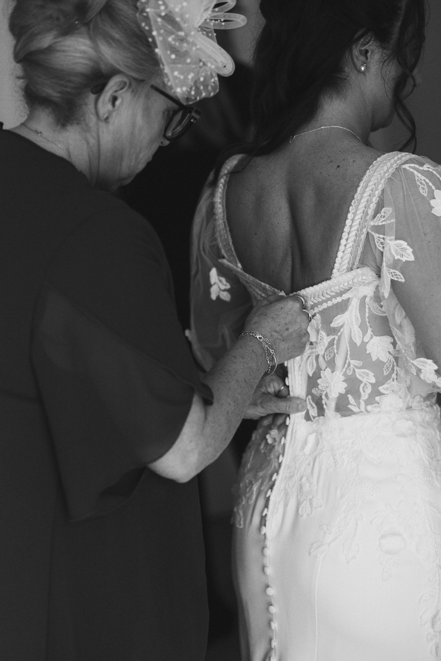Black and white photo of a woman helping a bride lace up her floral wedding dress from behind.