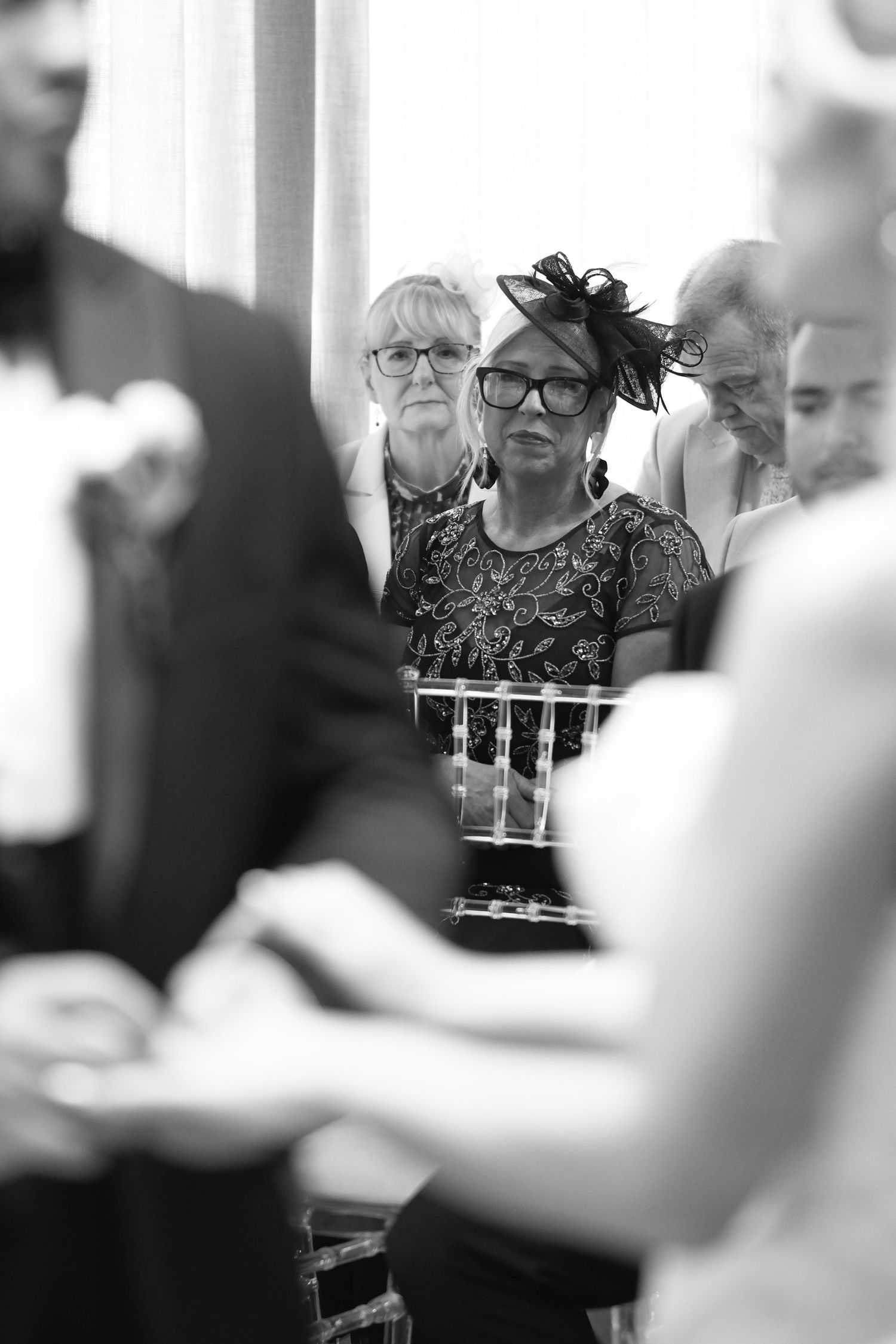Black and white photo of wedding guests watching ceremony, woman in glasses and fascinator hat in focus.