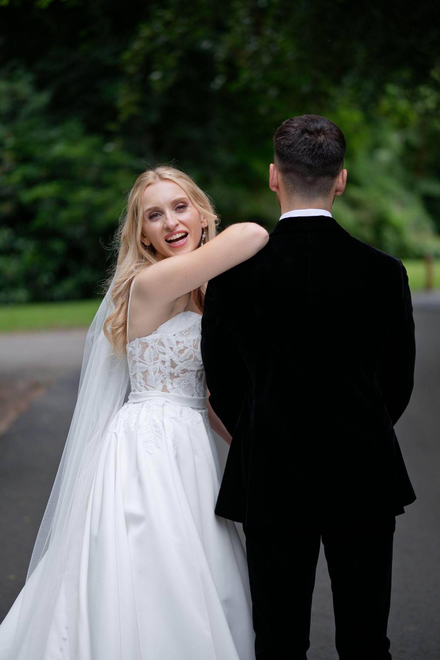 Smiling bride in white lace gown with veil embraces groom in black tuxedo outdoors with green trees.