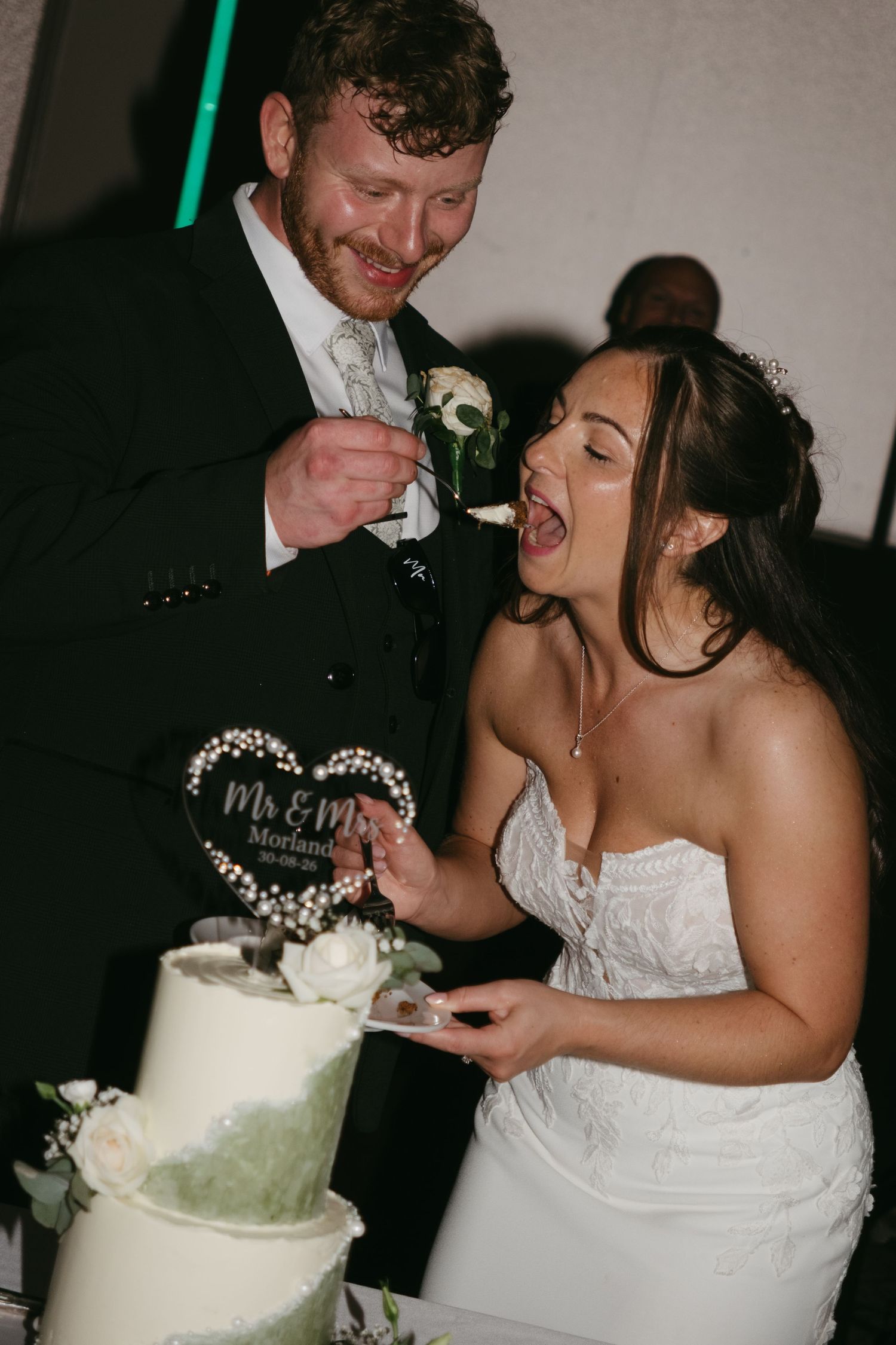Bride and groom joyfully cutting their white wedding cake topped with a Mr & Mrs heart topper at their wedding reception.