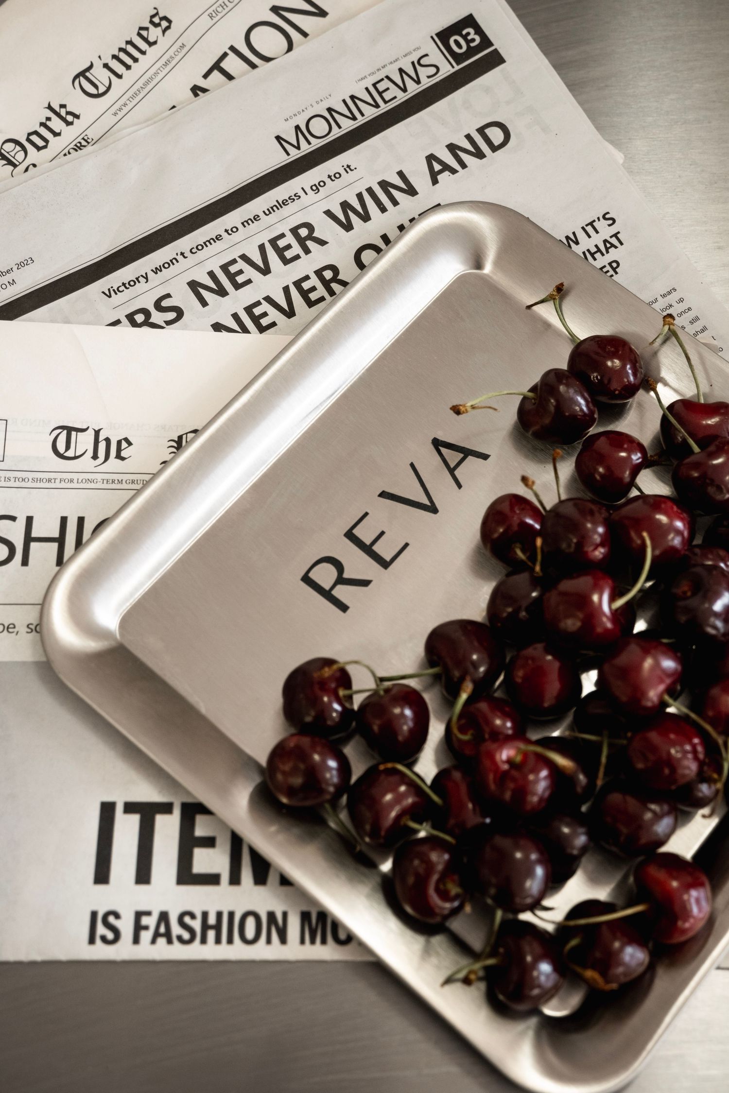 REVA branded silver tray filled with fresh dark cherries placed on fashion newspapers including New York Times.