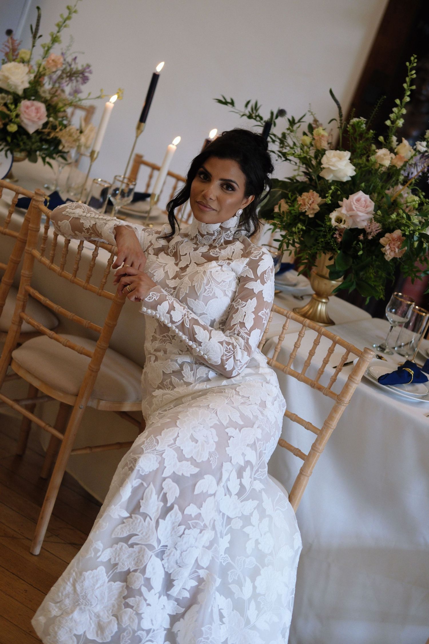 Bride in long-sleeve lace wedding gown seated at elegantly decorated reception table with floral centerpieces.