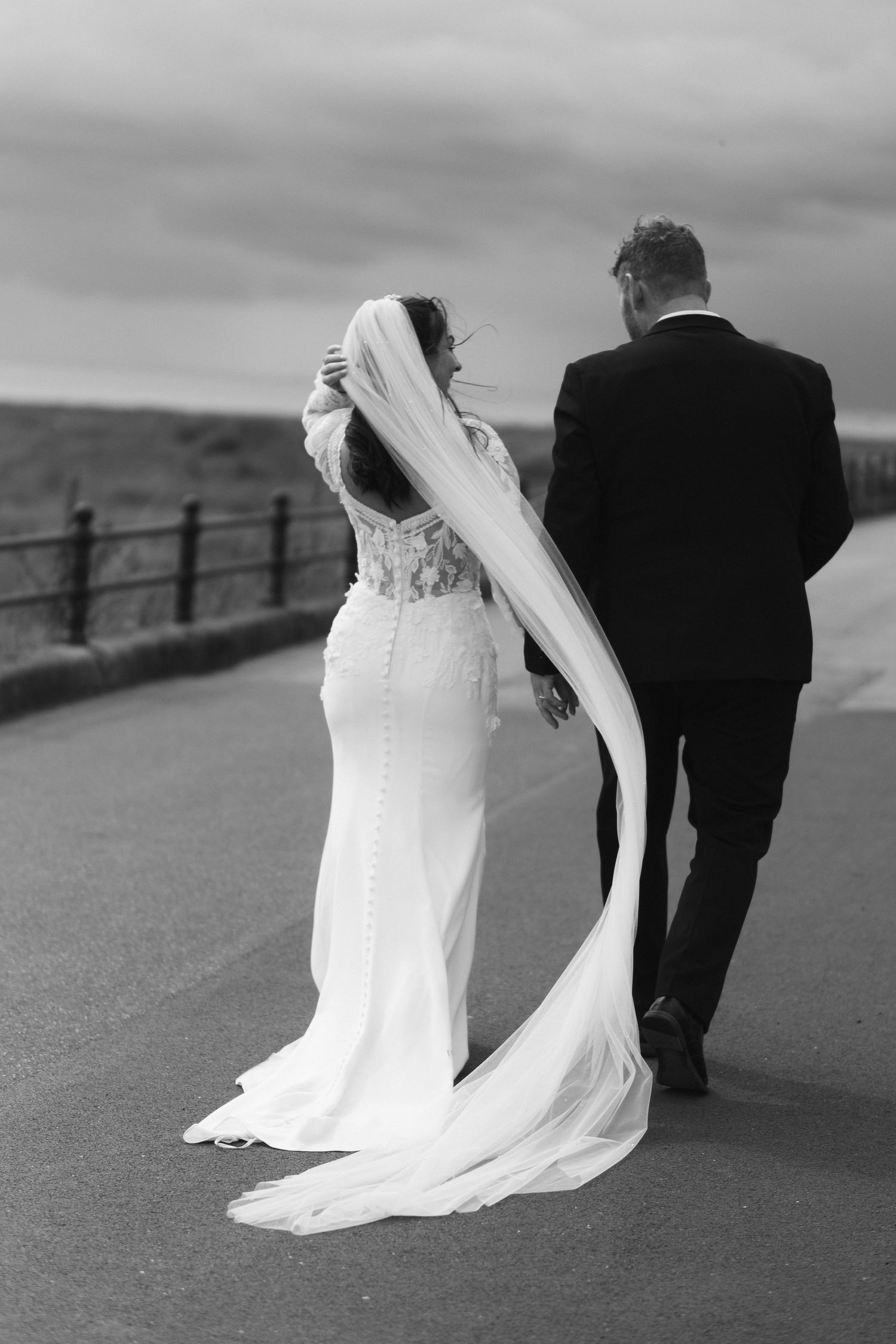 Black and white photo of bride and groom walking away on a road, brides veil flowing in the wind.