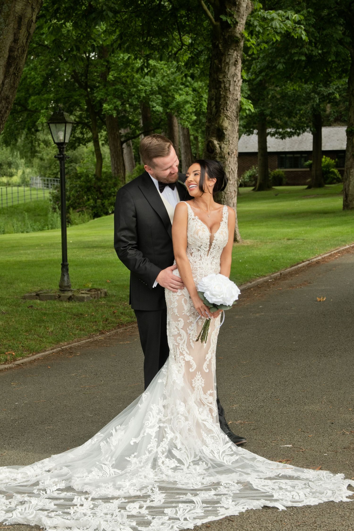 Groom in black tuxedo embraces smiling bride in lace wedding gown holding white bouquet on scenic garden path.