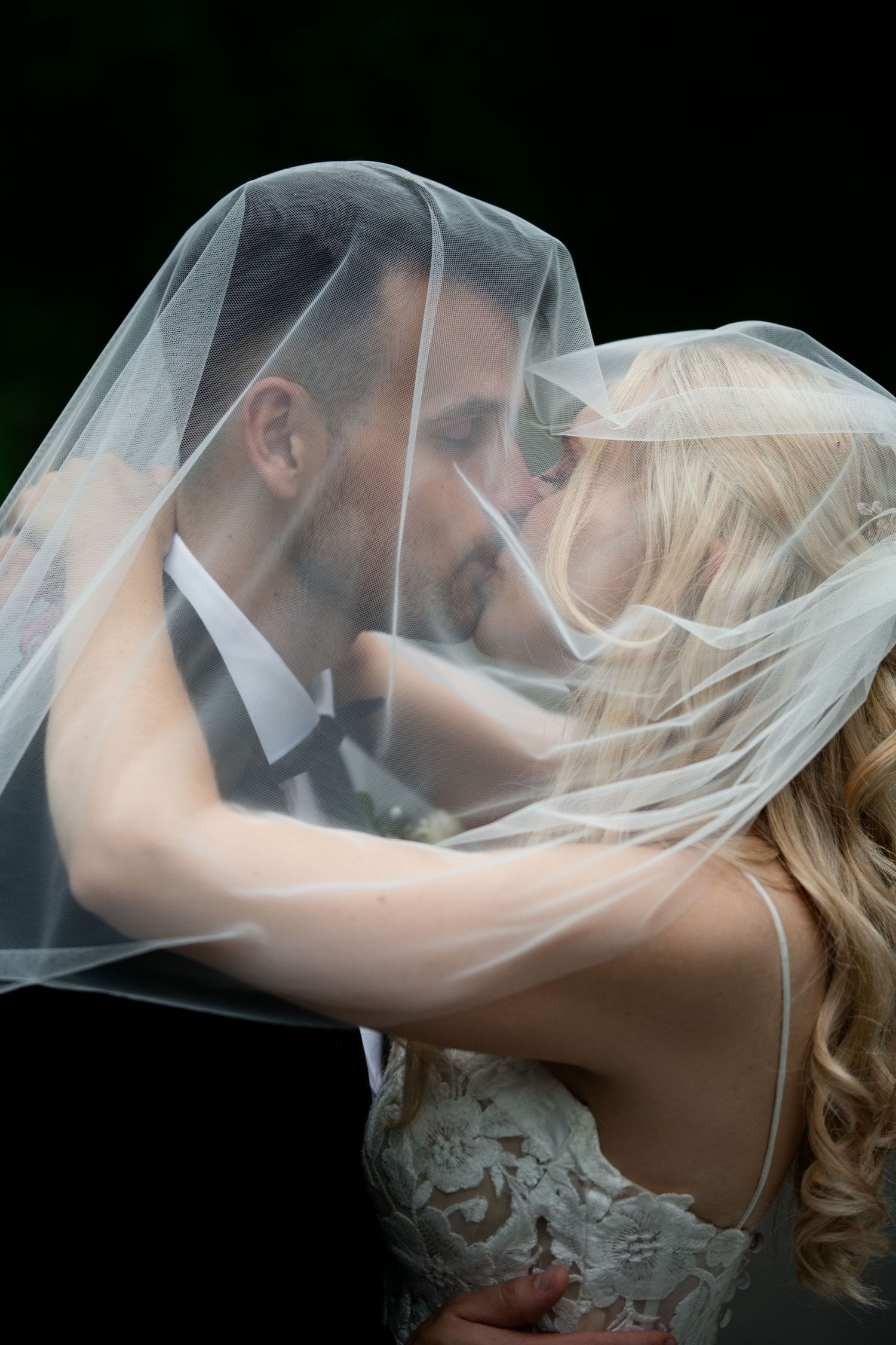 Romantic bride and groom sharing a kiss beneath flowing white wedding veil against dark background.