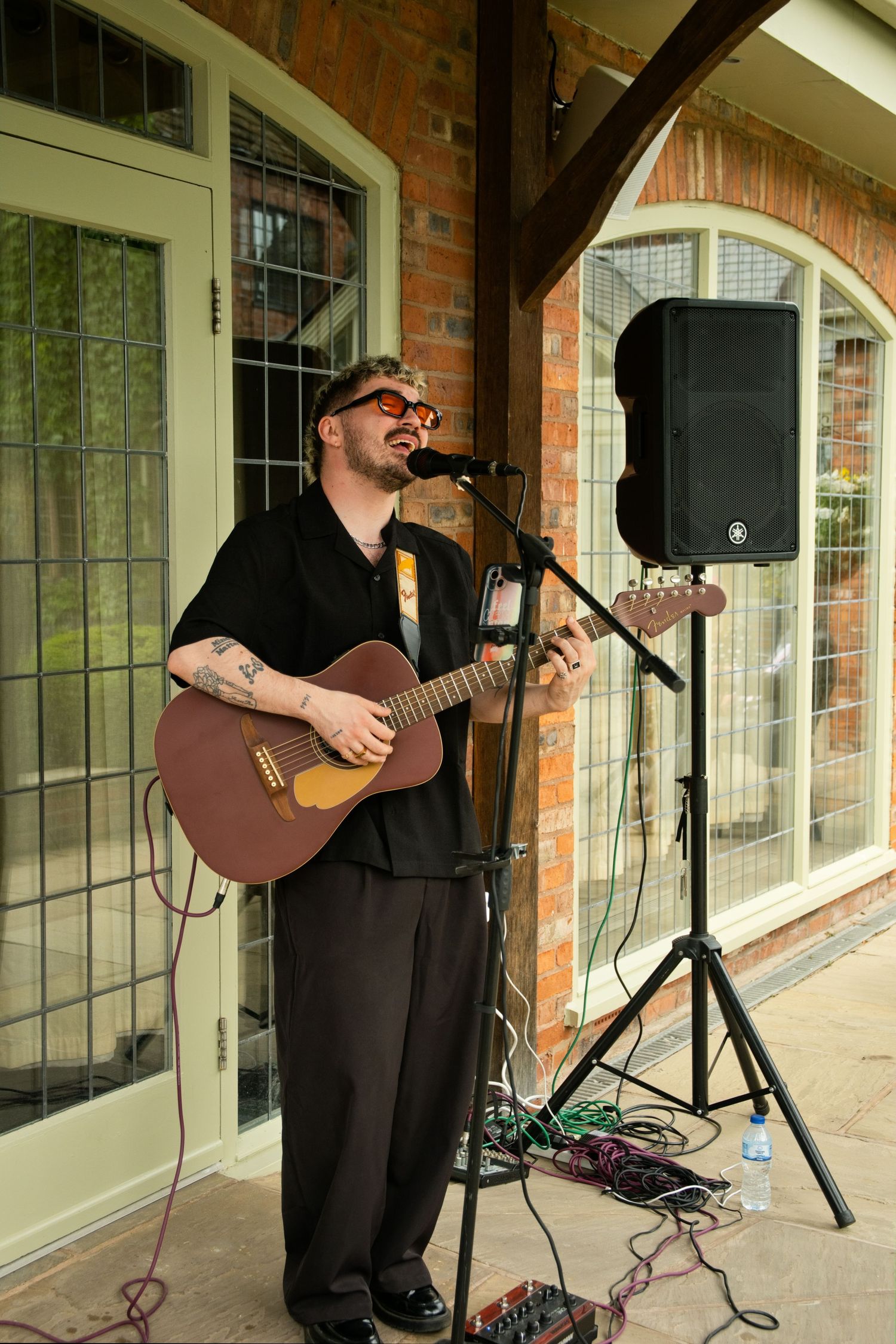 Tattooed musician playing acoustic guitar and singing into microphone at outdoor wedding reception.