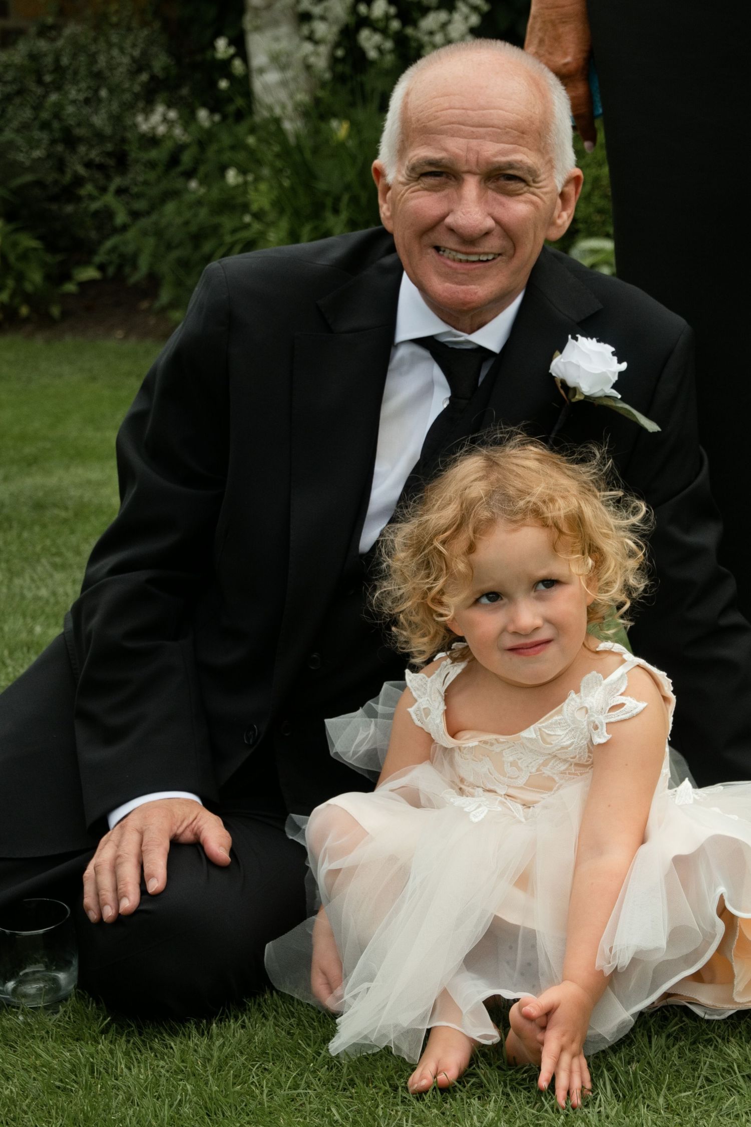 Elderly gentleman in black tuxedo smiling while posing with adorable curly-haired flower girl on grass.