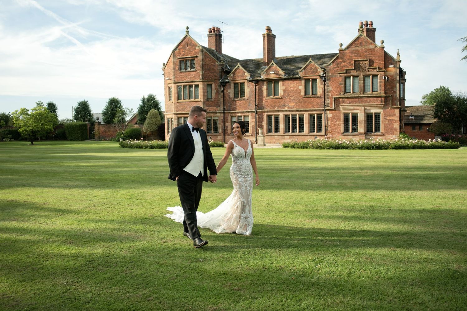 Newlyweds walking hand-in-hand on lawn before grand manor house.