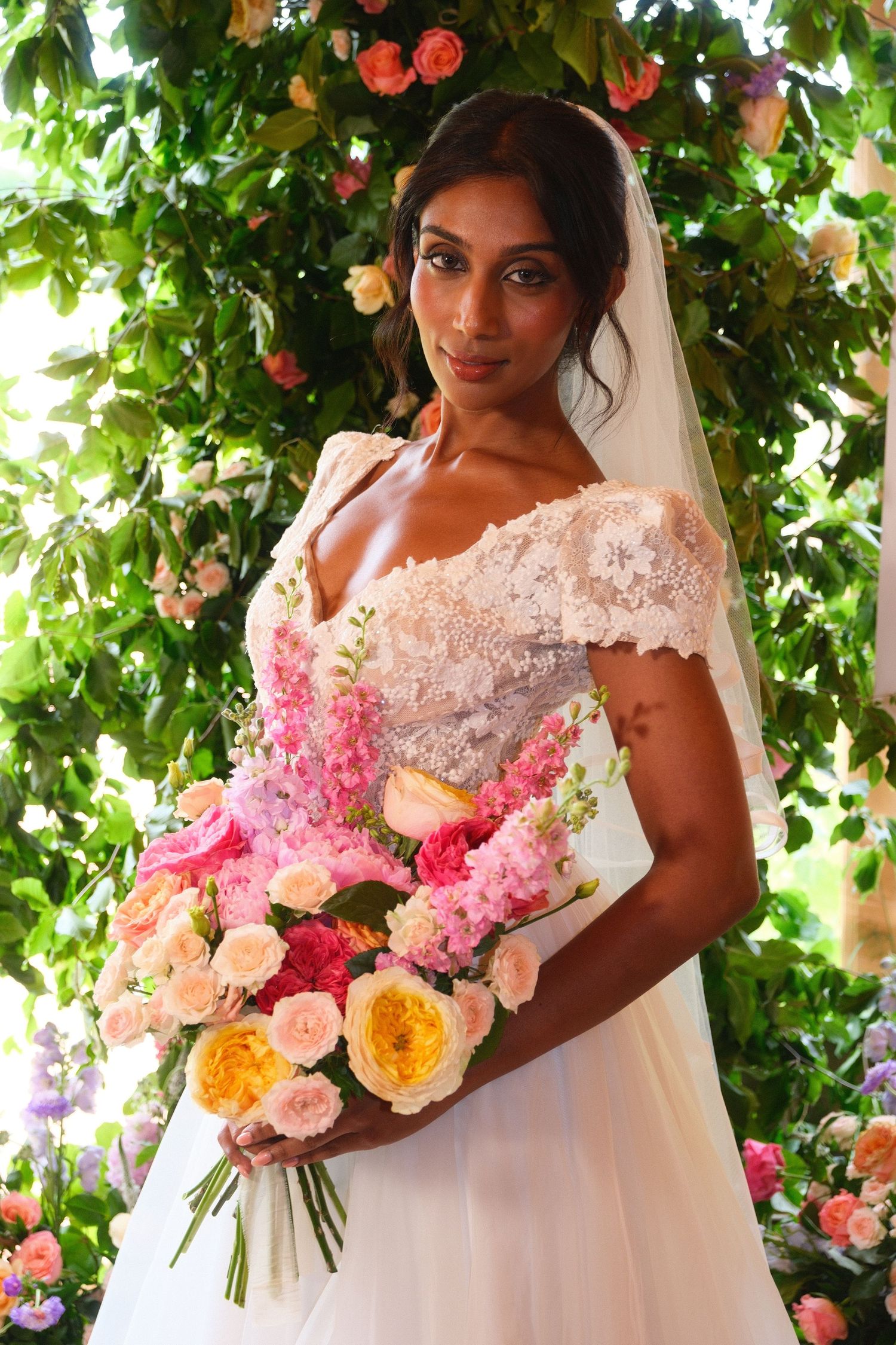 Beautiful bride in lace off-shoulder gown holding vibrant pink and yellow floral bouquet.