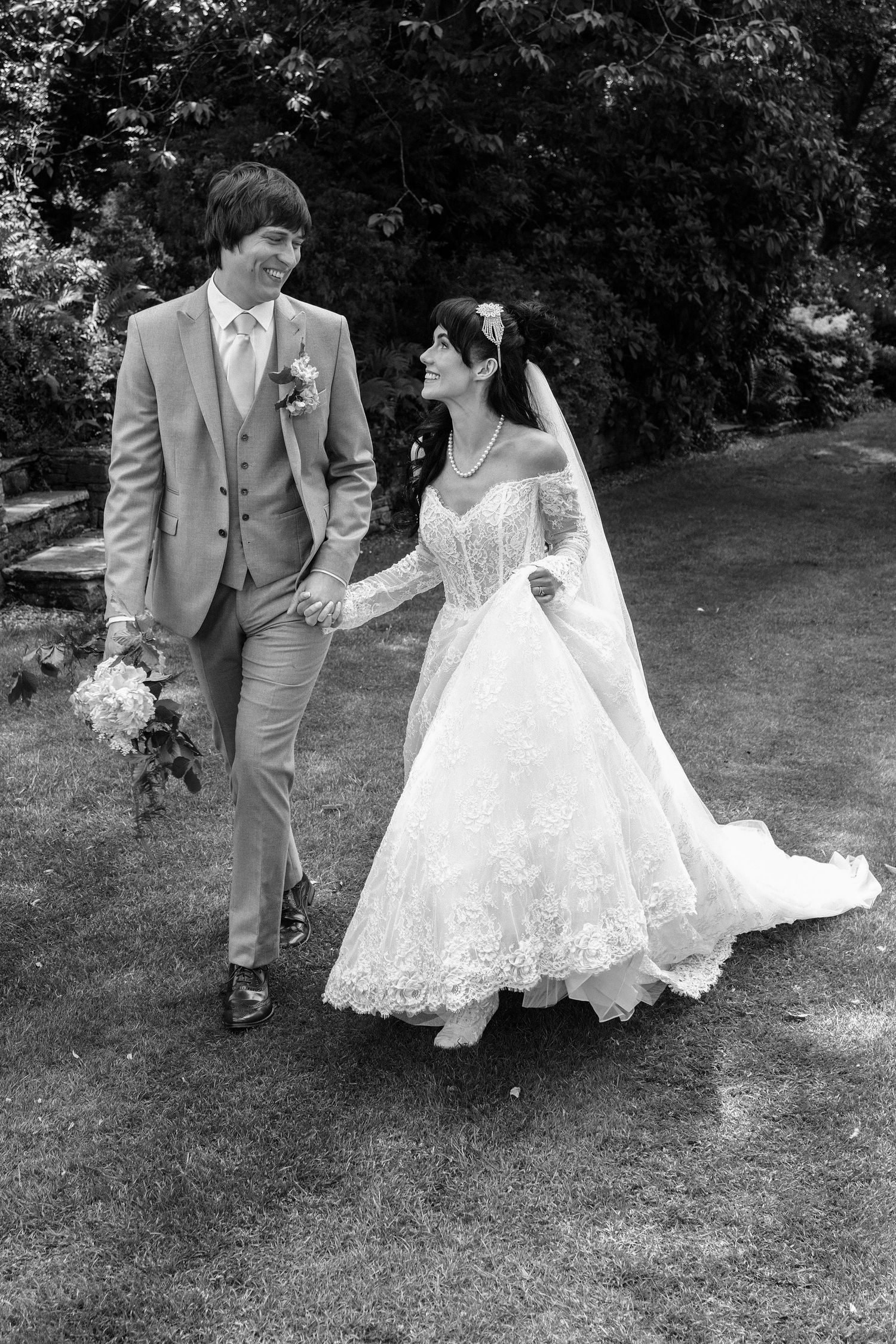 Black and white photo of a smiling bride and groom holding hands, walking through a garden on their wedding day.