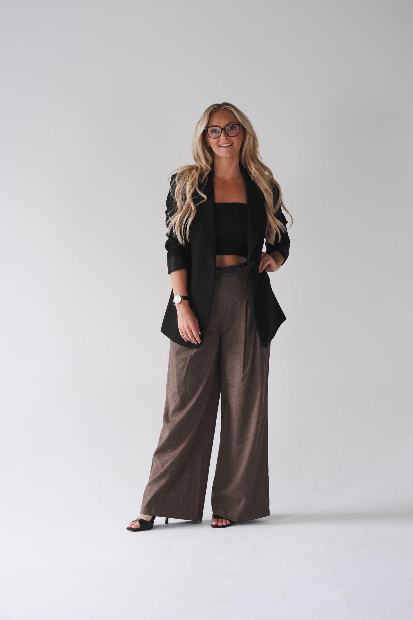 Woman in black blazer, crop top, and wide-leg brown pants with heels and glasses on white background.