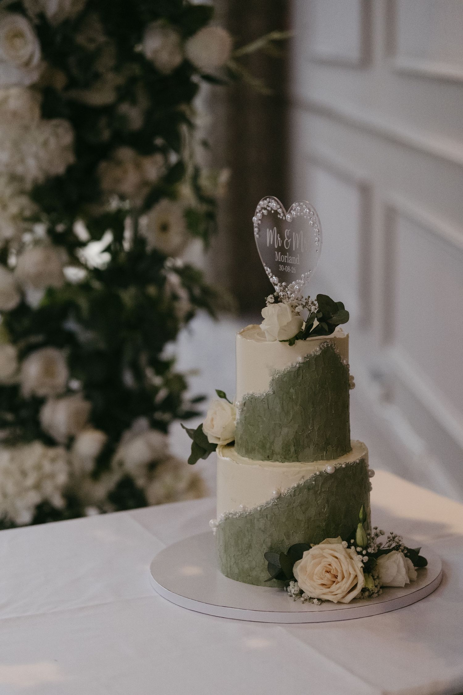 Two-tier white and sage green wedding cake with roses and heart topper, displayed before a white floral backdrop.