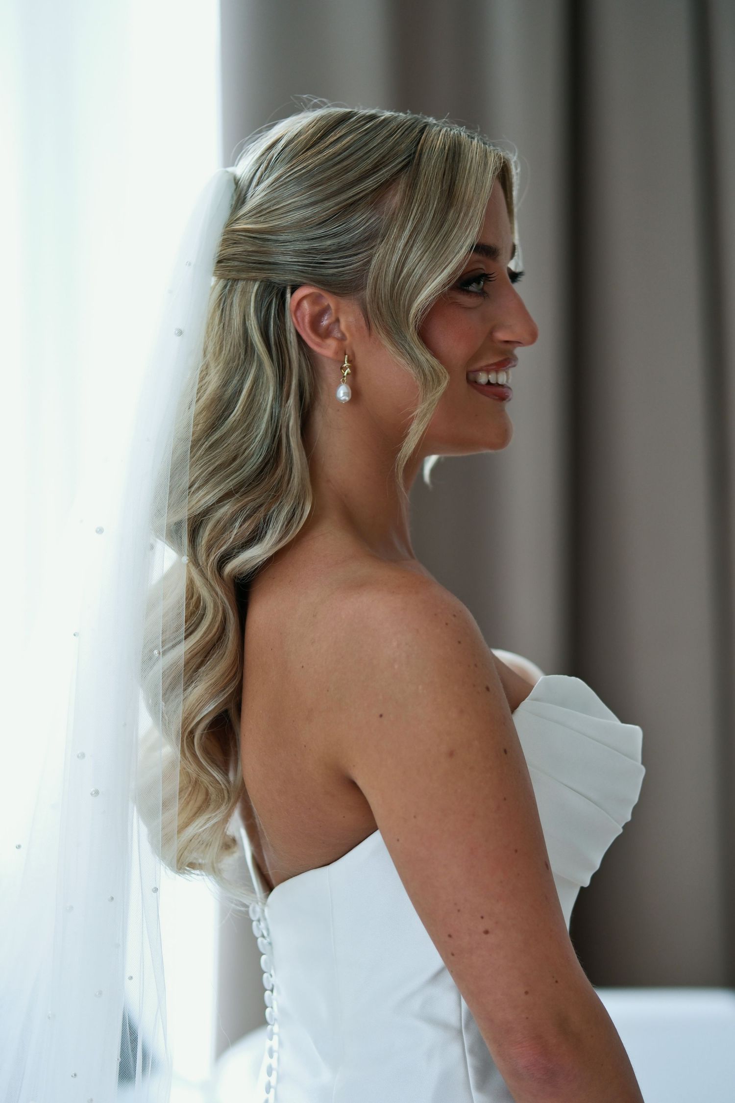 Radiant bride in strapless white gown with veil, pearl earrings, and soft blonde waves smiling by window.