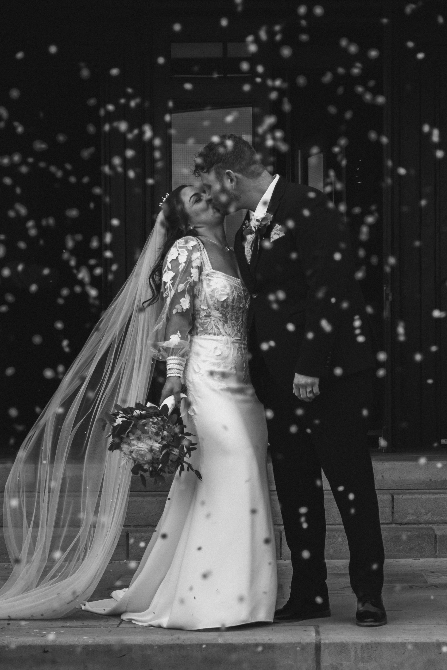 Black and white photo of a bride and groom sharing a kiss on church steps, surrounded by falling confetti.