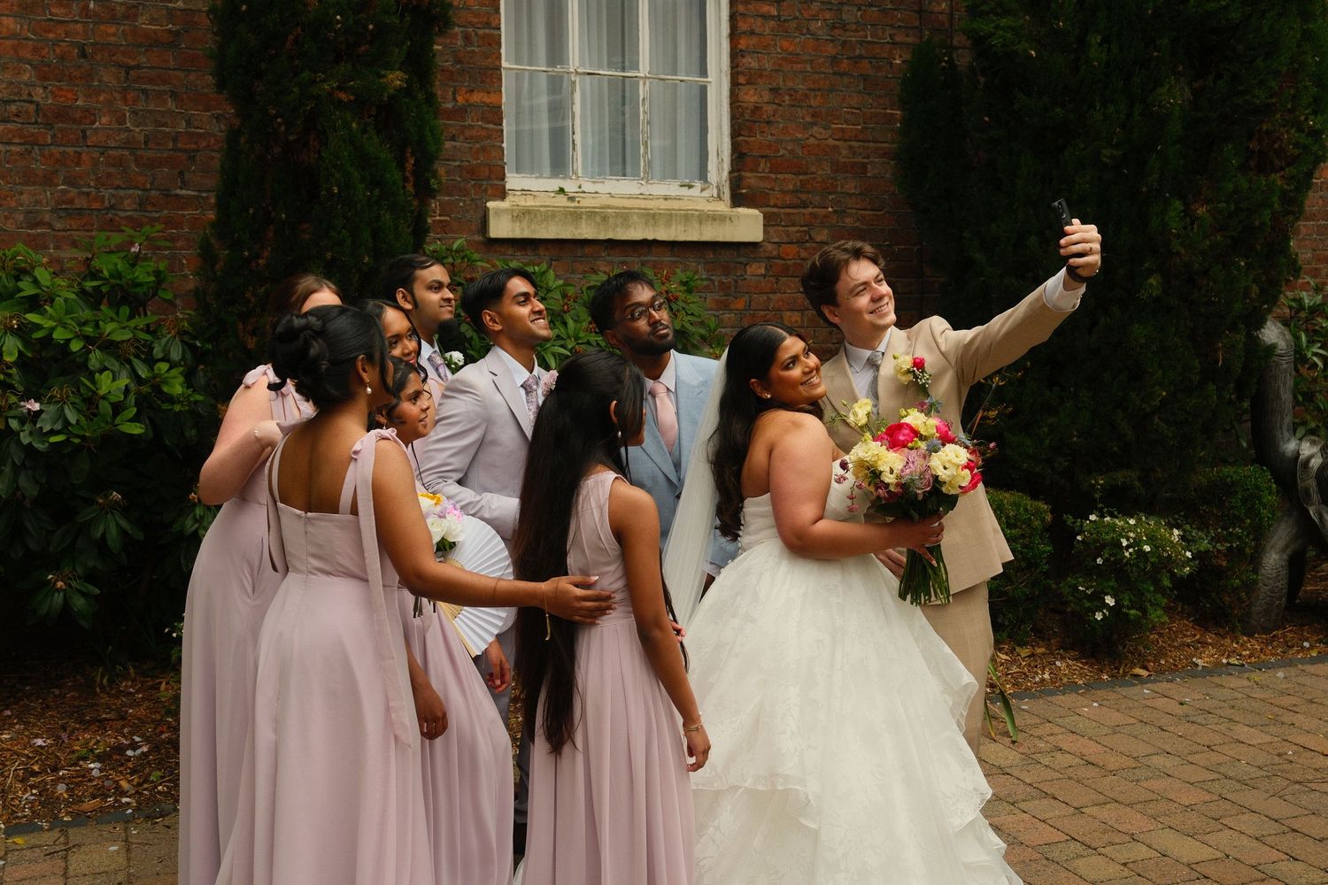 Wedding party takes a group selfie outdoors, bridesmaids in mauve dresses with bride holding colorful bouquet.