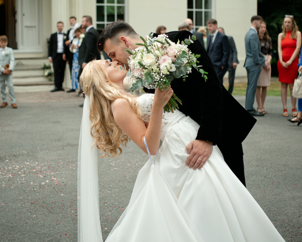 Couple sharing a romantic dip kiss outside their wedding venue while guests cheer behind them.