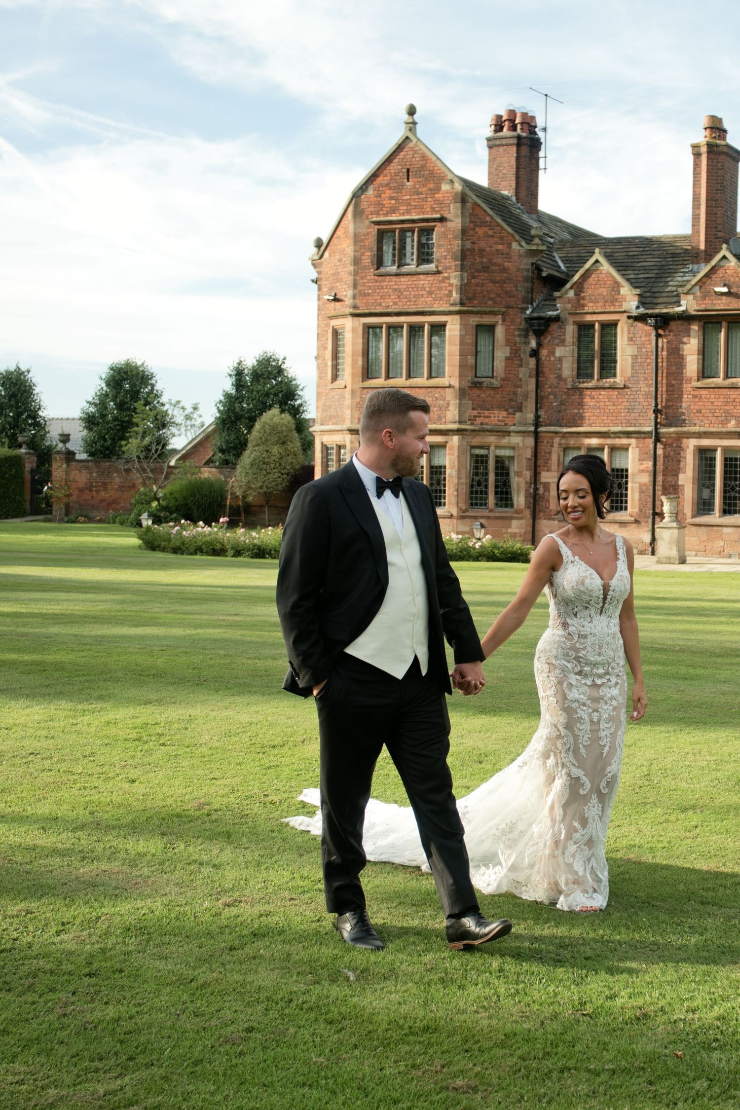 Bride and groom walk hand in hand across lawn before grand red brick English manor house on wedding day.