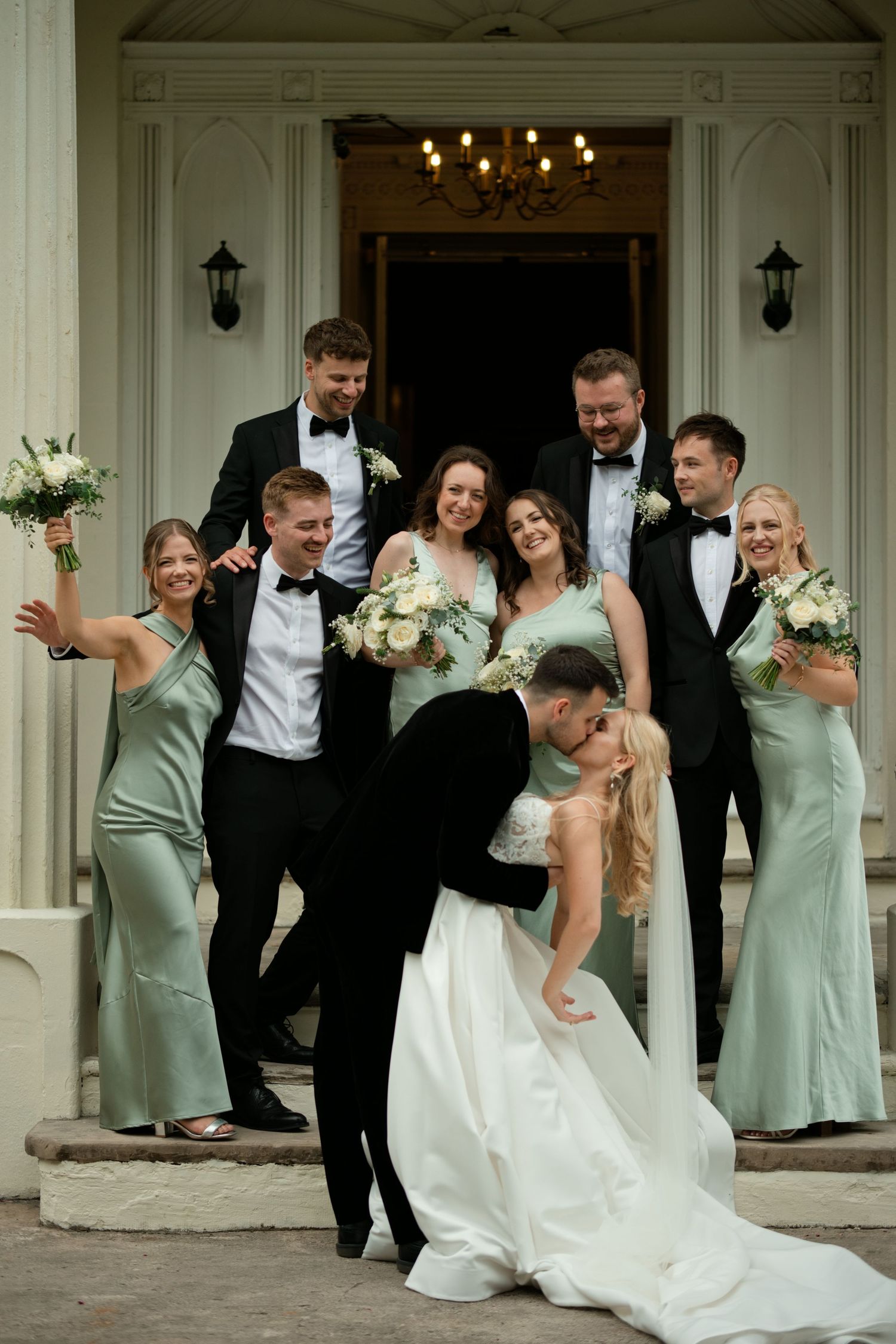 Wedding party in black tuxedos and sage green bridesmaid dresses celebrating as groom dips bride on steps.