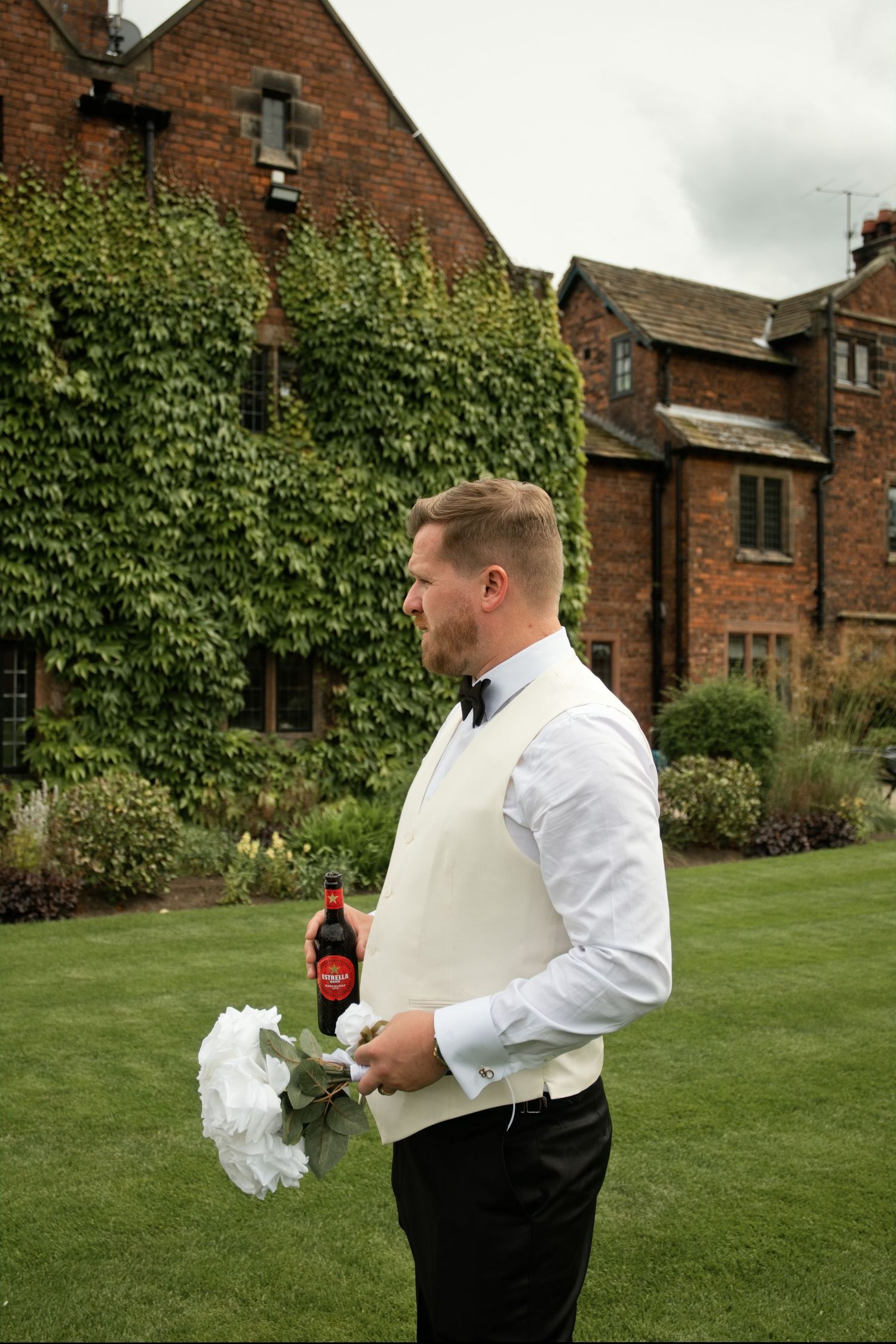 Groom in white vest holding white bouquet and Coca-Cola bottle in front of ivy-covered manor.