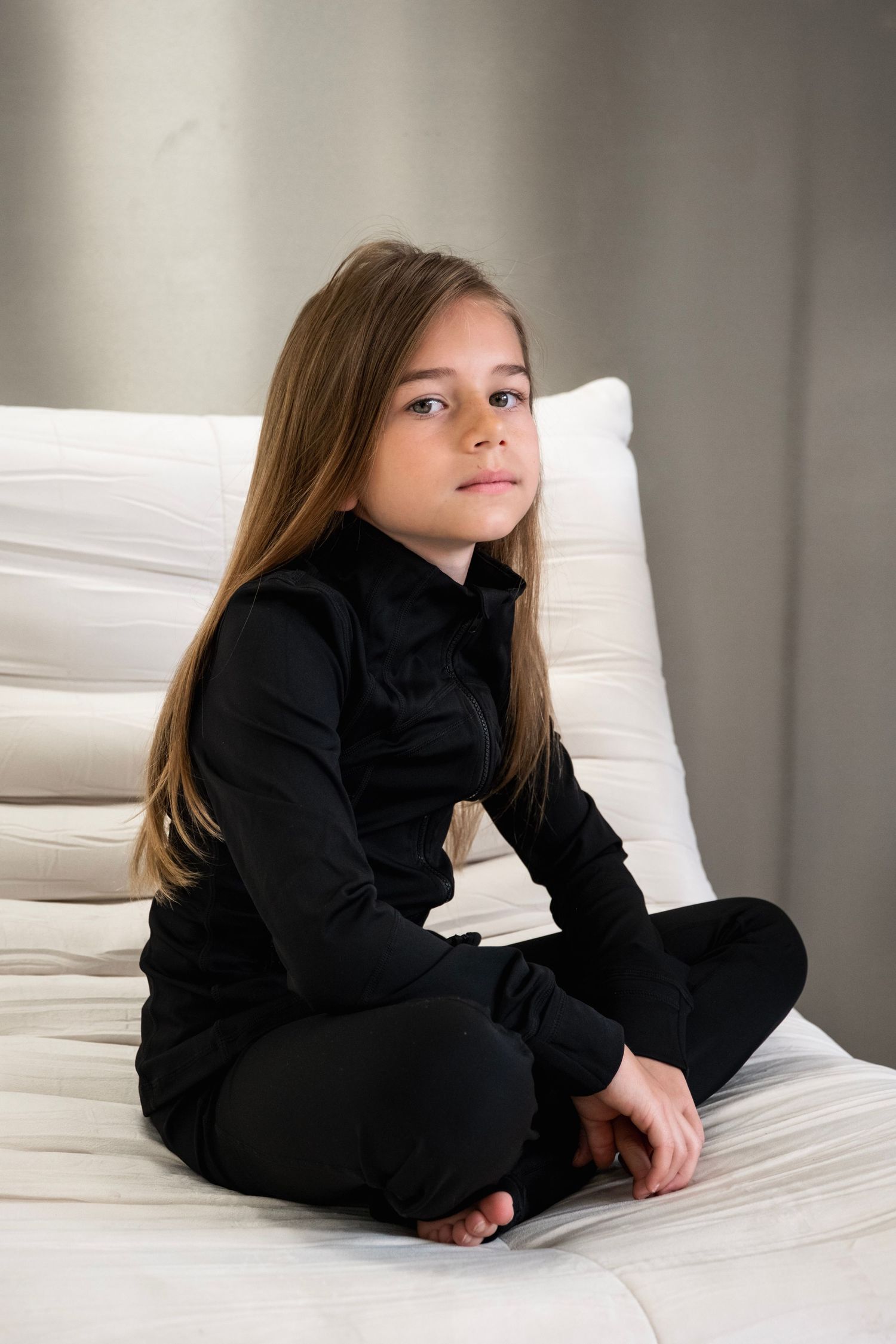 A young girl with long hair sits cross-legged on a white bed, wearing an all-black outfit, looking calmly at the camera.