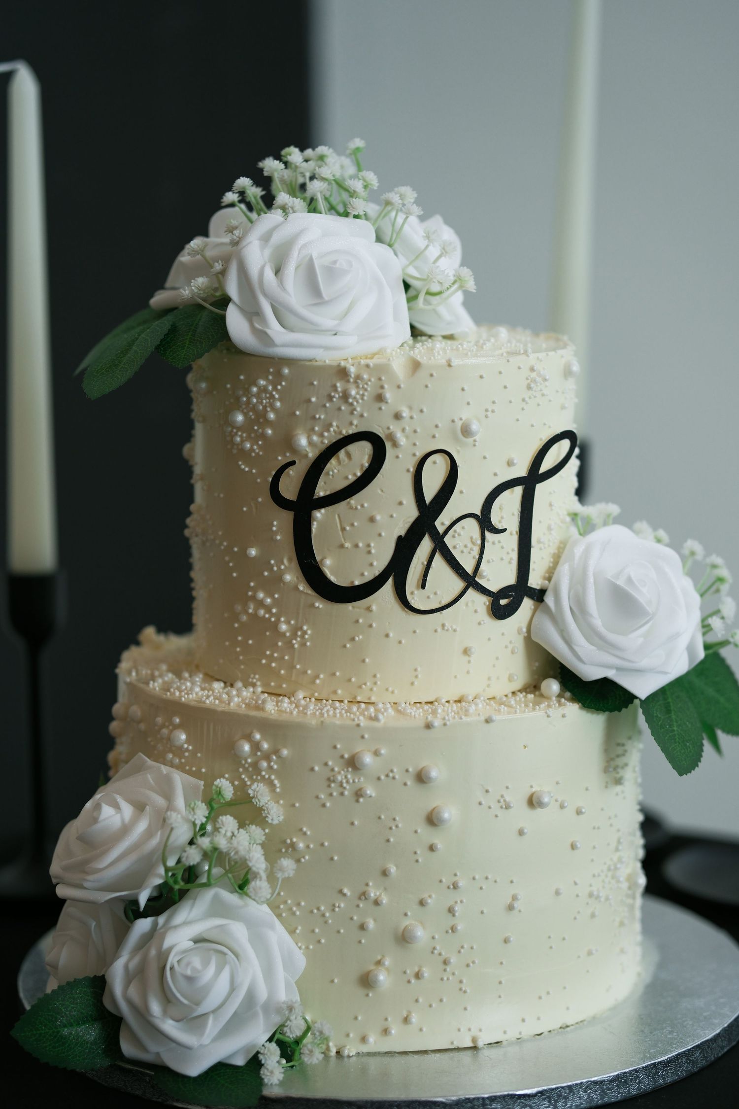 Two-tier white wedding cake with pearl details, white roses, and black monogram C&L topper on silver stand.