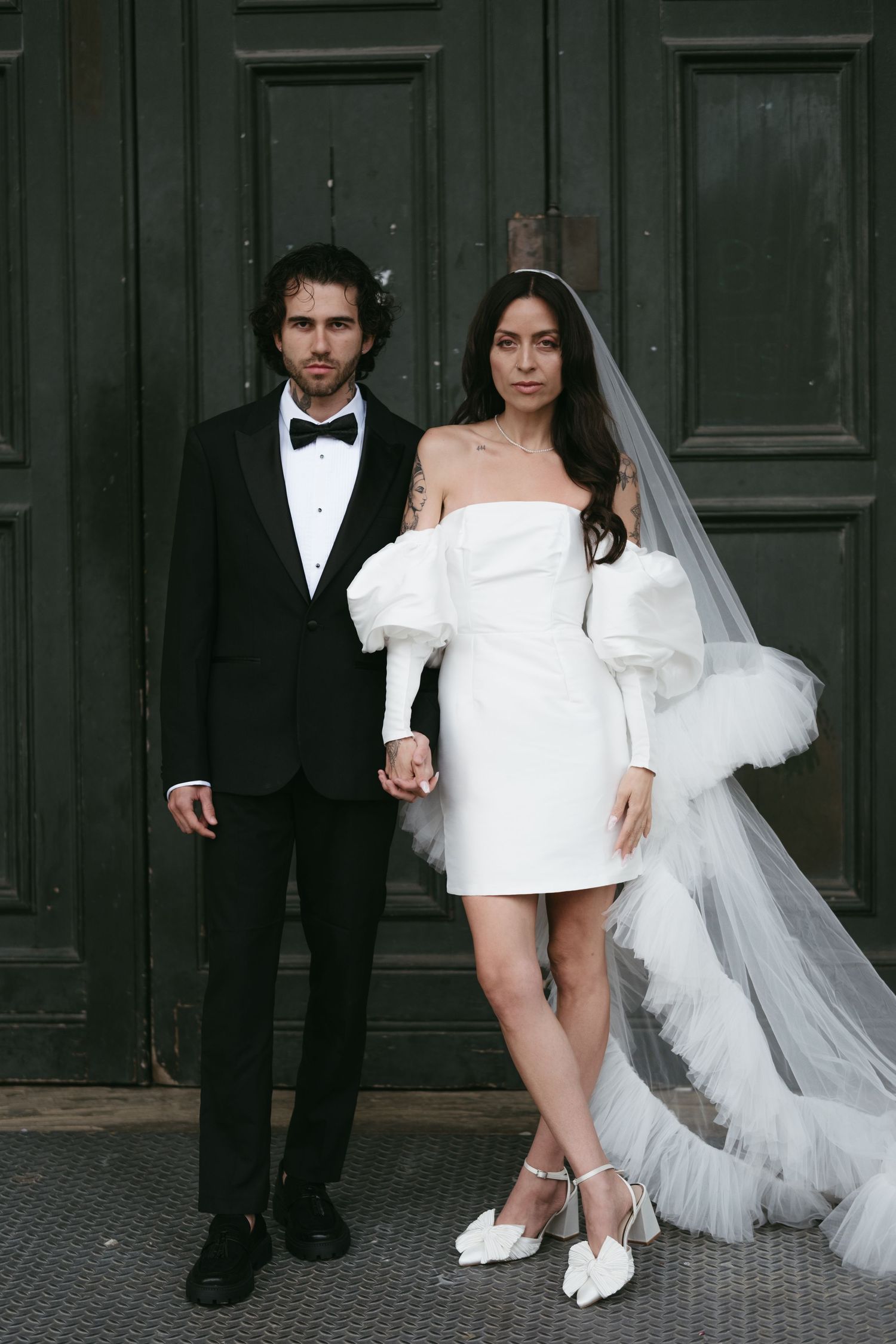 Bride in white off-shoulder mini dress with veil and groom in black tuxedo posing before dark wooden doors.