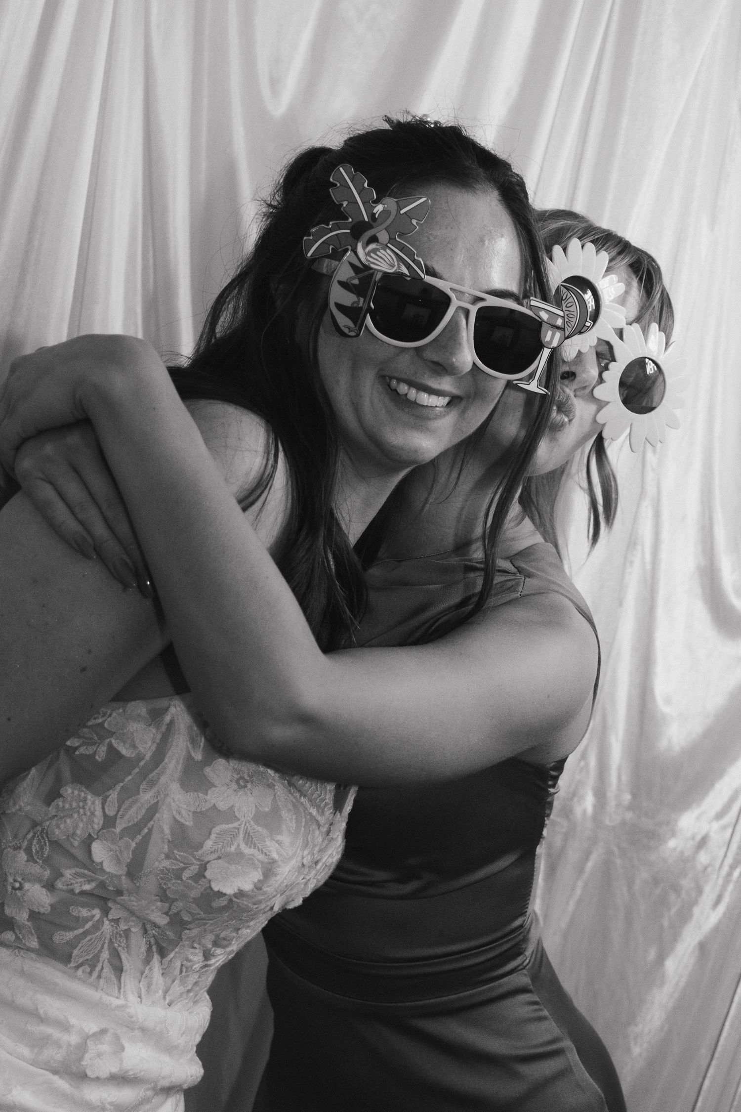 Two women hugging and smiling, wearing fun sunglasses and hair accessories, in a black and white photo.