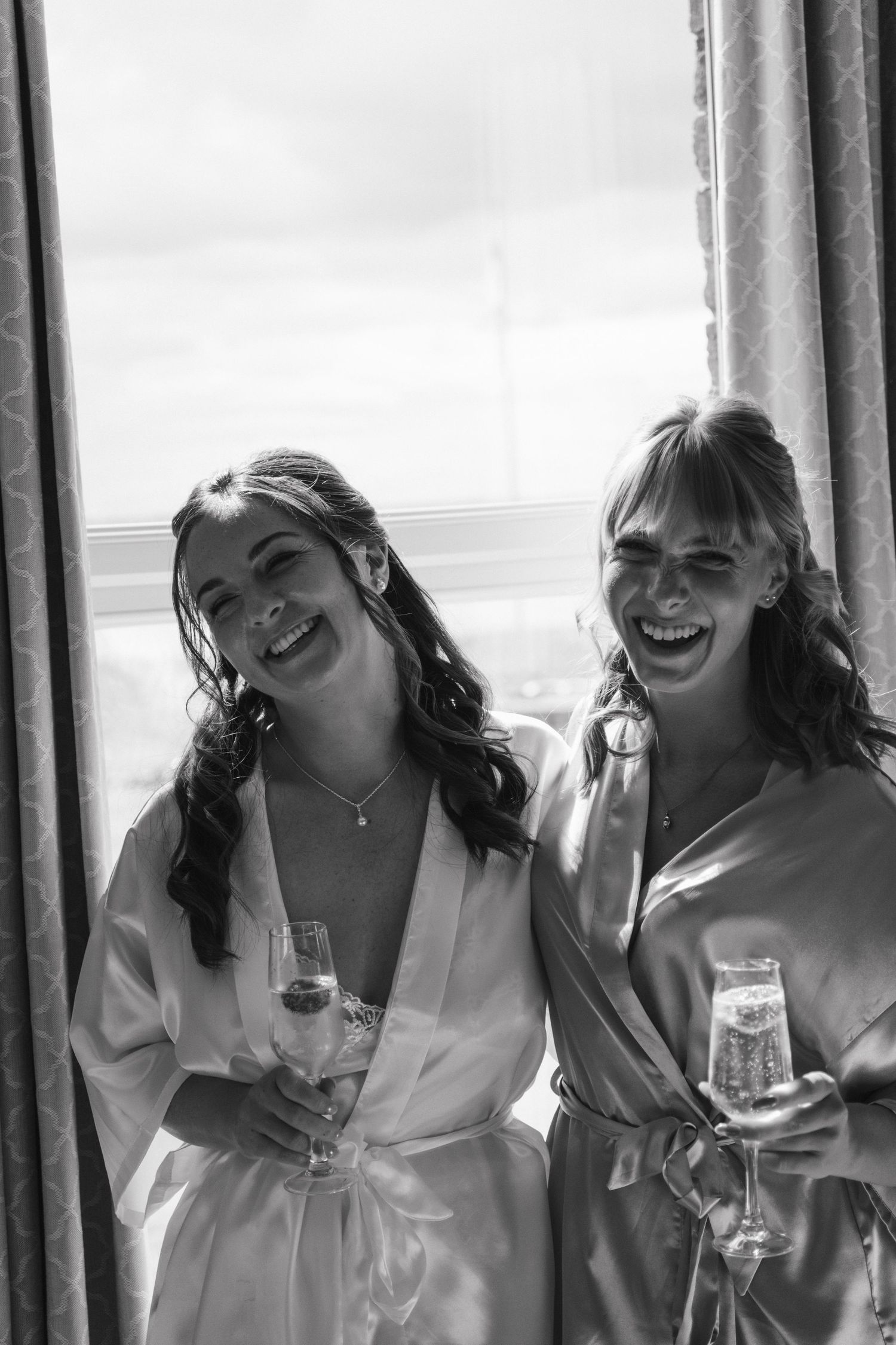 Two laughing bridesmaids in satin robes holding champagne glasses standing by a window in black and white photo.