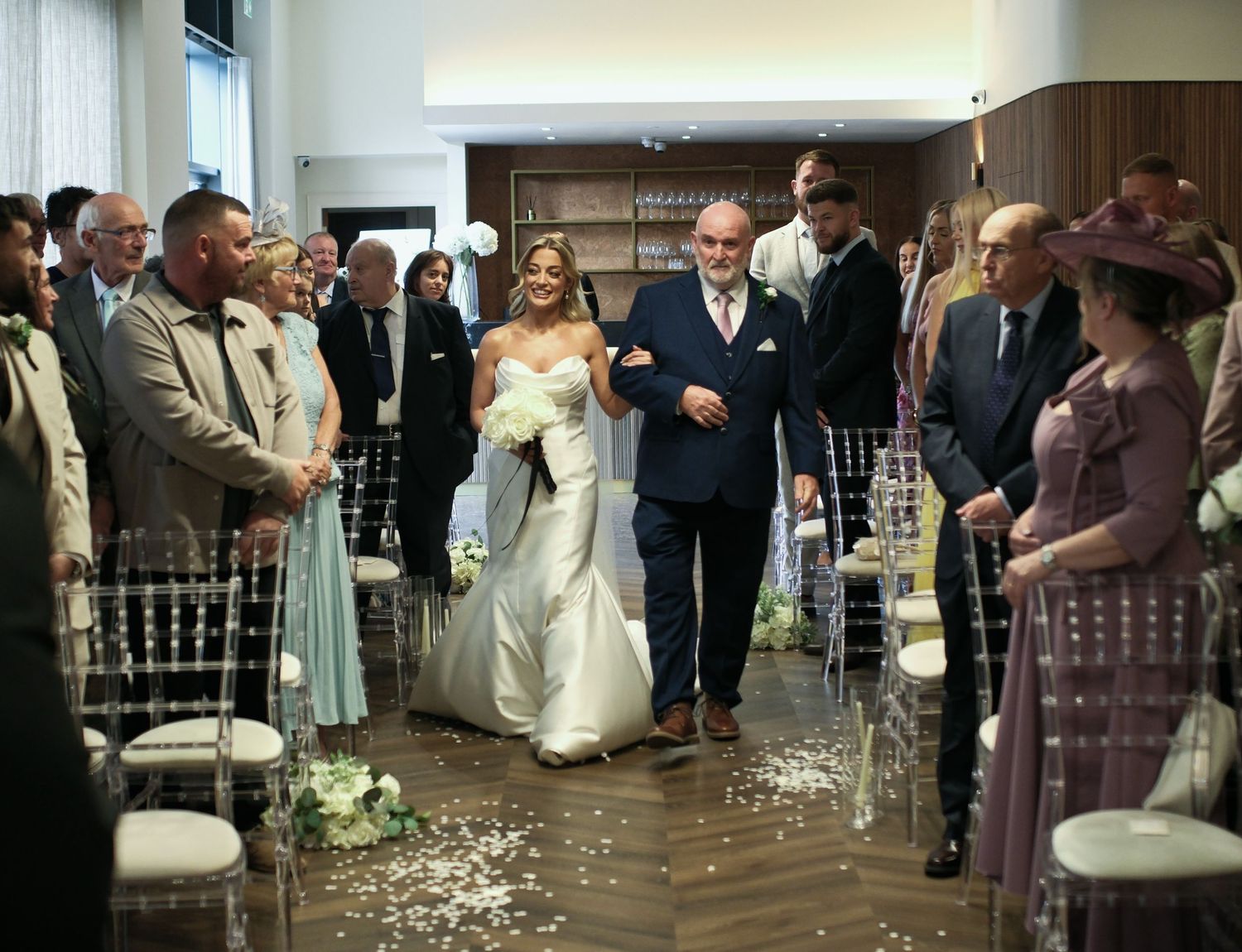 Bride walking down the aisle escorted by her father in a modern wedding ceremony venue.