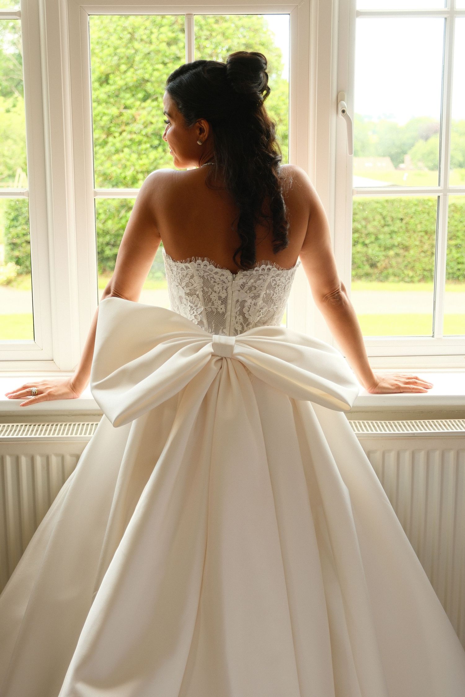 Bride in strapless lace corset gown with large satin bow gazing out a bright window.