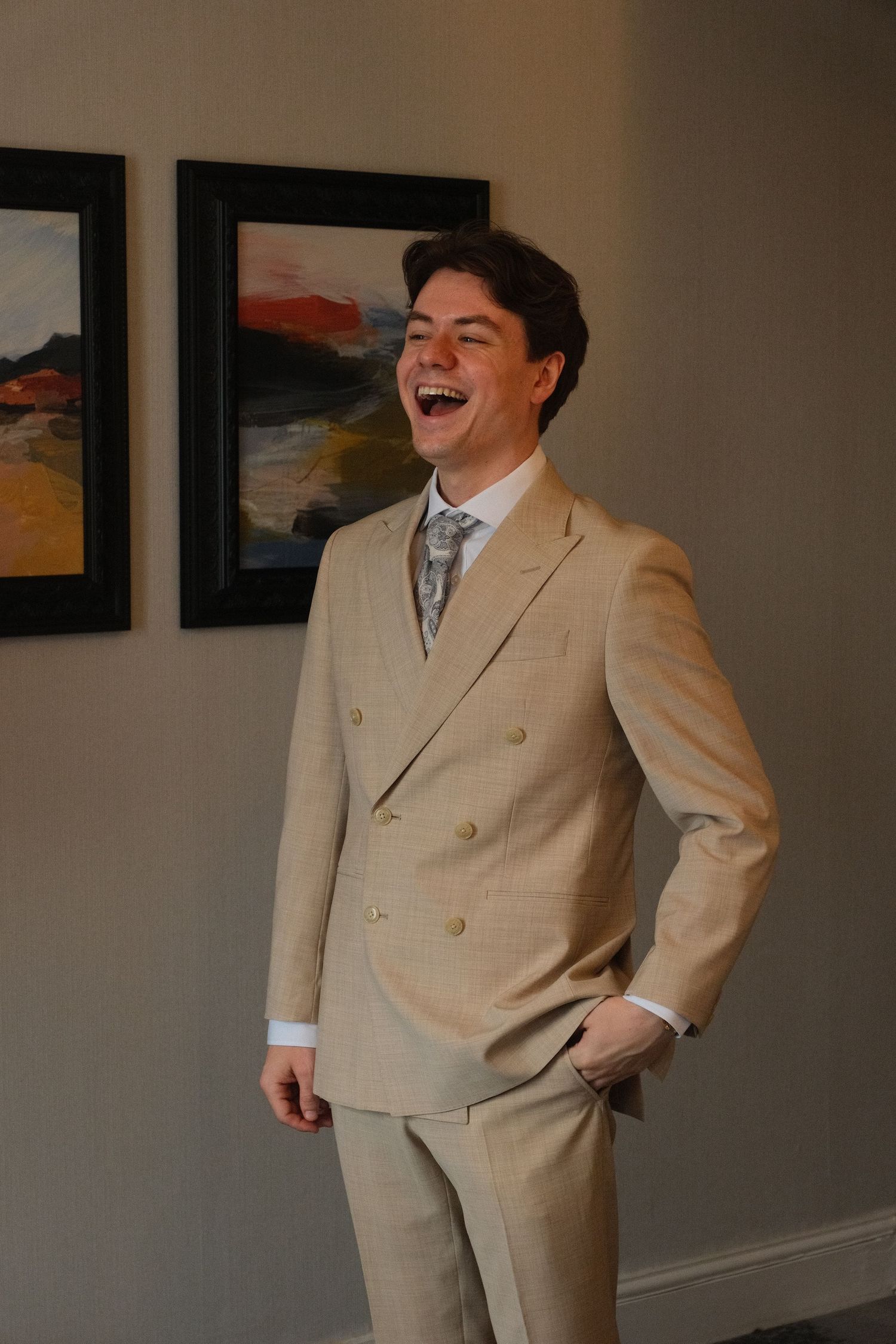 Laughing young groom in beige double-breasted suit stands in front of abstract wall art.
