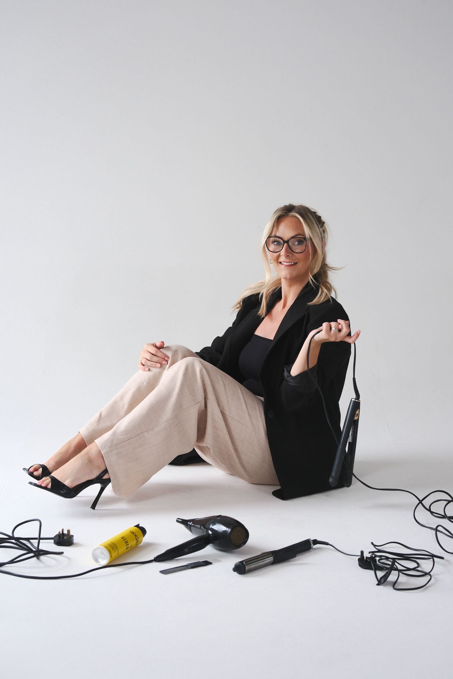 Woman in glasses and black blazer sitting on floor surrounded by hair styling tools and accessories on white background.