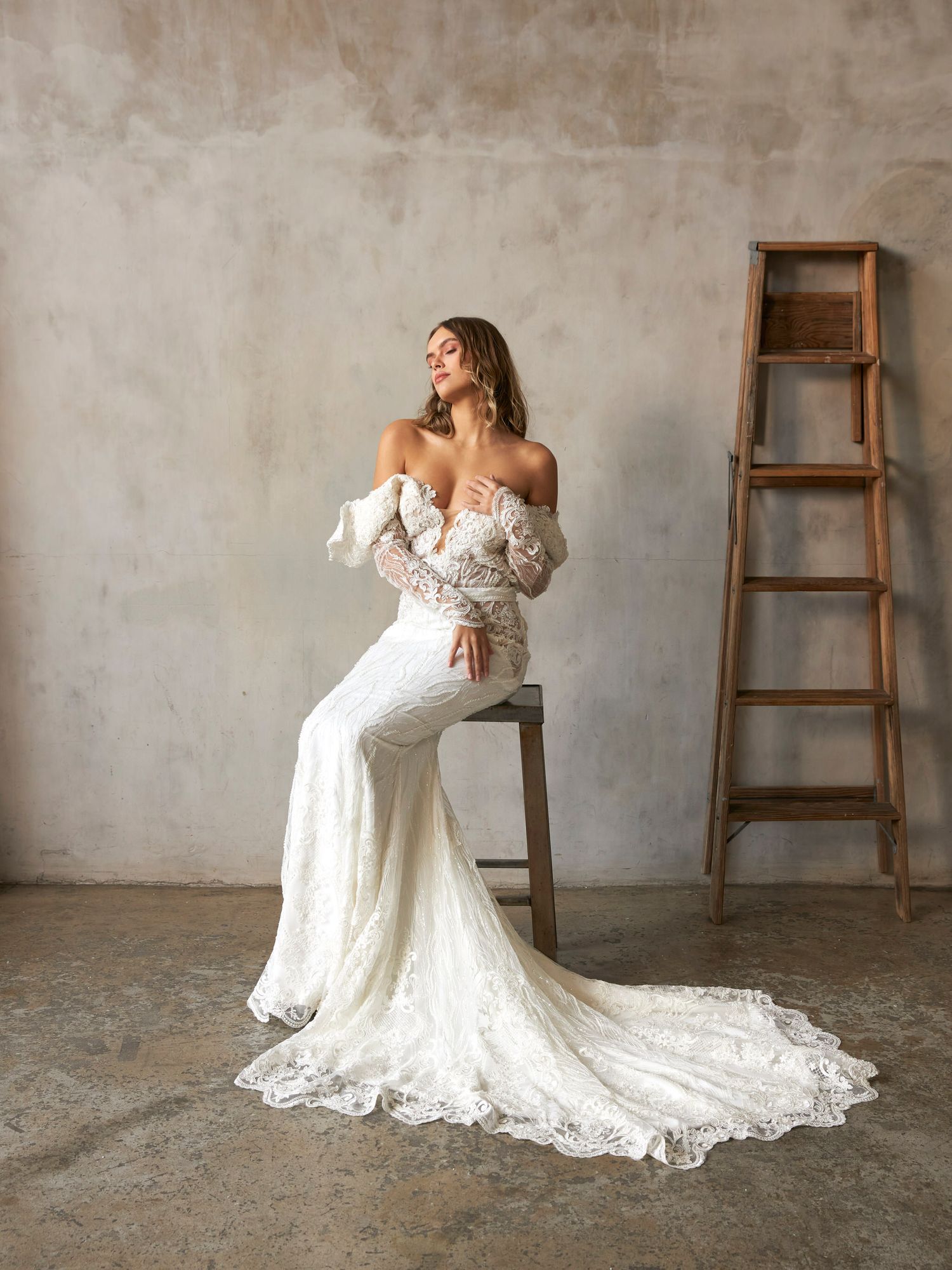 Bride in off-shoulder lace mermaid wedding gown with long train seated near rustic wooden ladder.