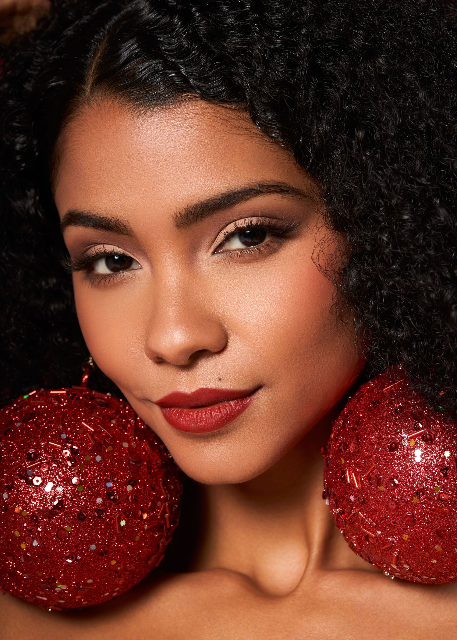 Beautiful woman with natural afro hair holding red glitter ornaments beside her face with red lipstick.