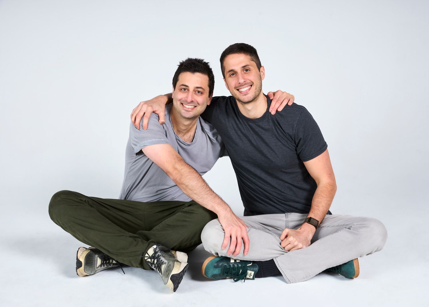 Two young men sit cross-legged on the floor smiling together in a casual studio portrait.