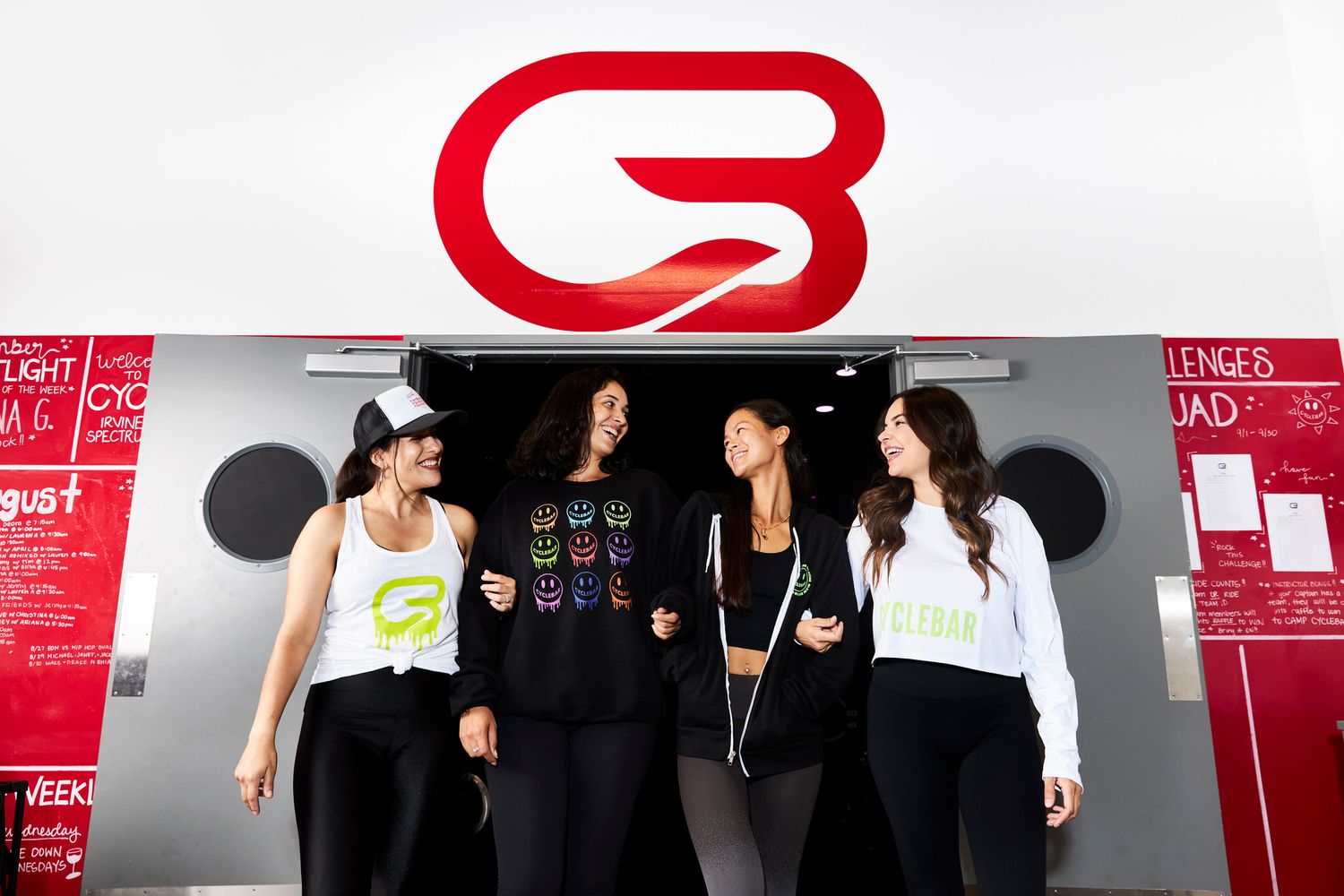 Four women in athletic wear laugh together outside a GB Fitness studio entrance.