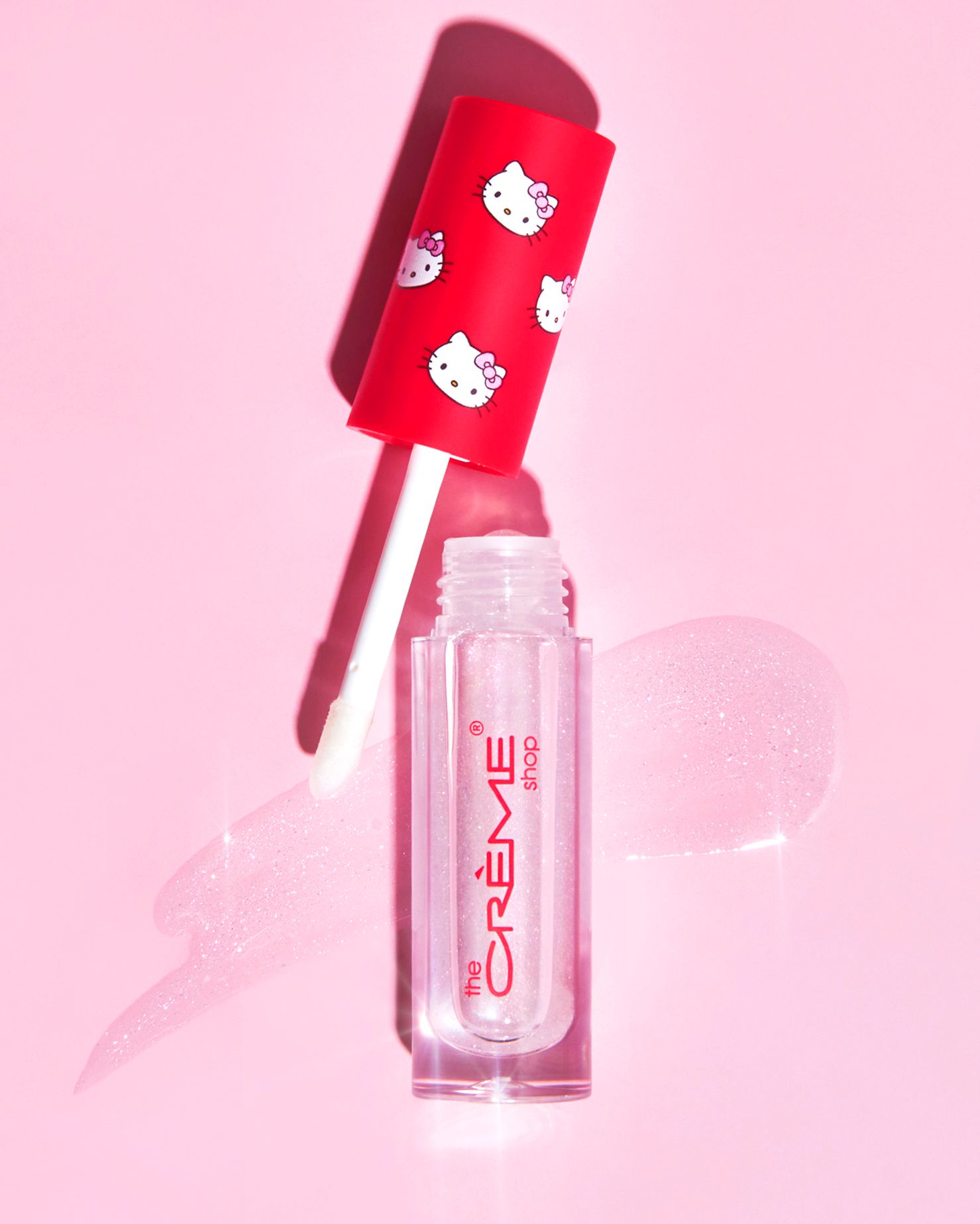 Hello Kitty x The Creme Shop clear lip gloss with red Hello Kitty printed applicator on pink background.