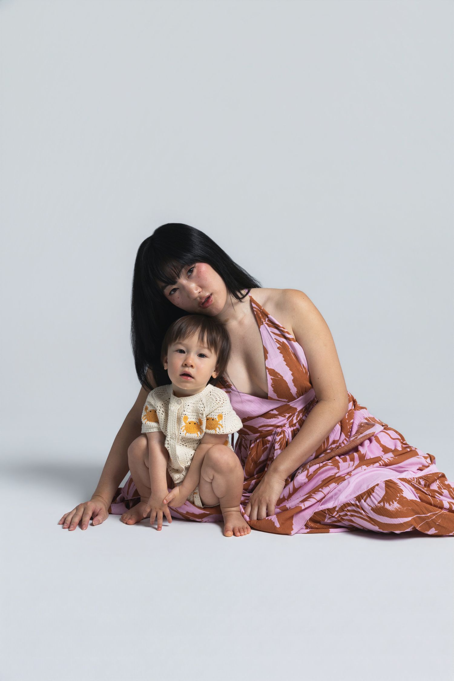 A woman in a pink floral dress sits on the floor with a toddler in a white outfit against a light gray background.