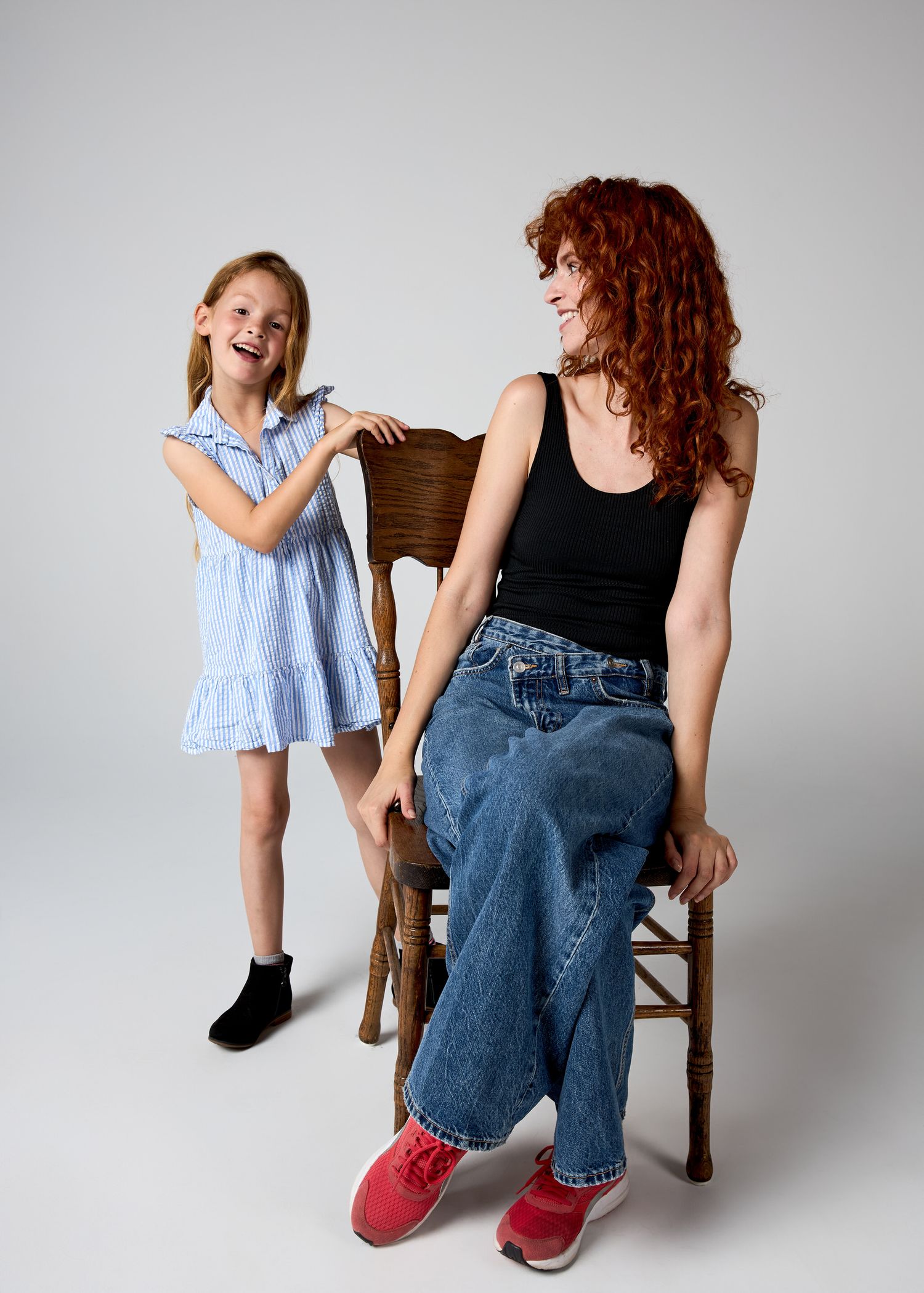 A red-haired woman sits on a wooden chair while a young blonde girl in a blue dress stands beside her, both smiling.