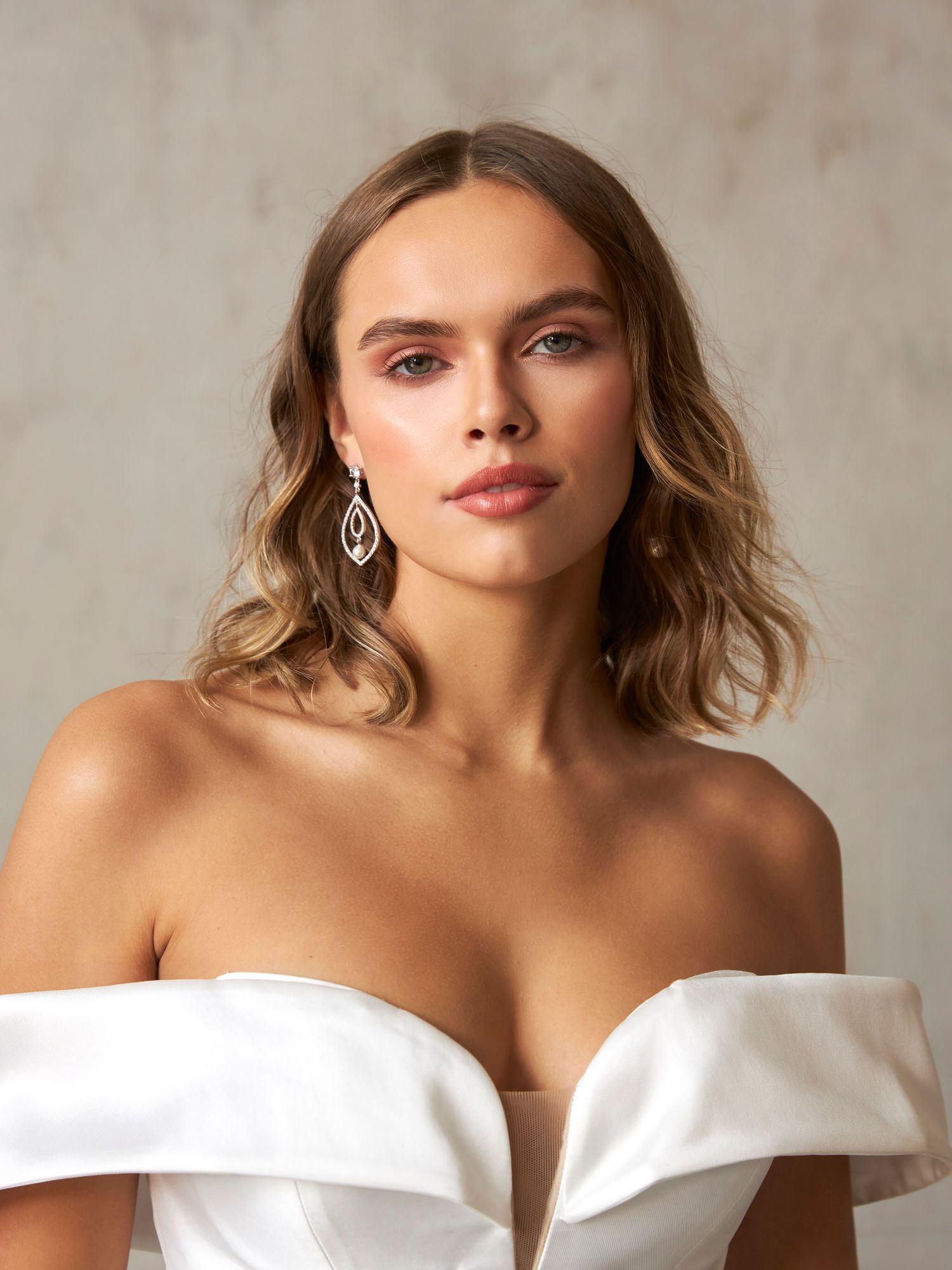 Elegant woman in off-shoulder white bridal gown wearing crystal drop earrings with soft waves.