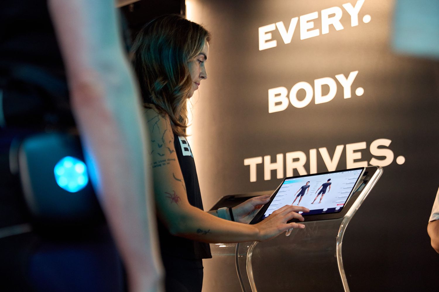 Woman interacts with a fitness body analysis touchscreen display under an 'Every Body Thrives' motivational wall sign.