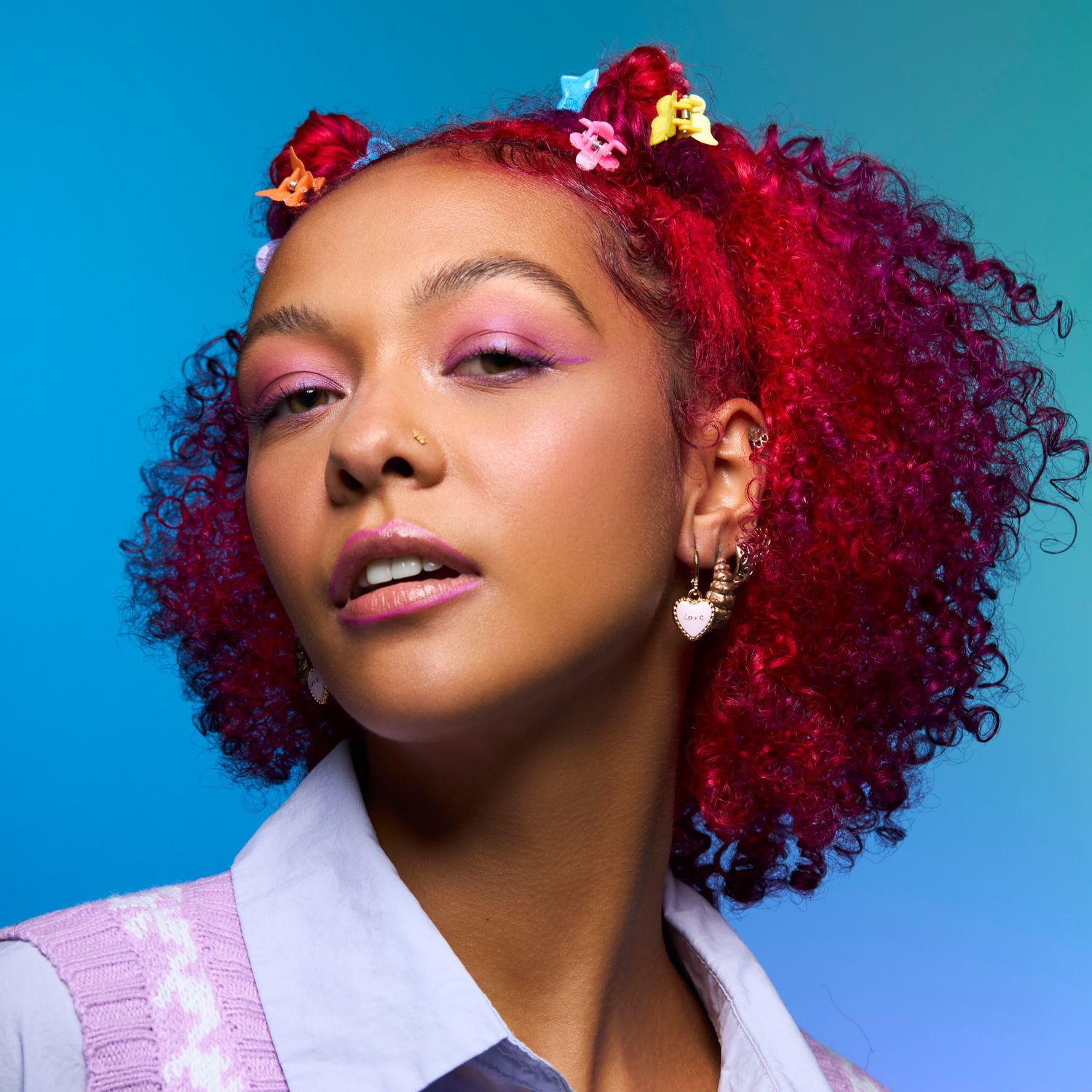 Model with vibrant red curly hair styled with colorful clips wearing pink eyeshadow and heart earrings.