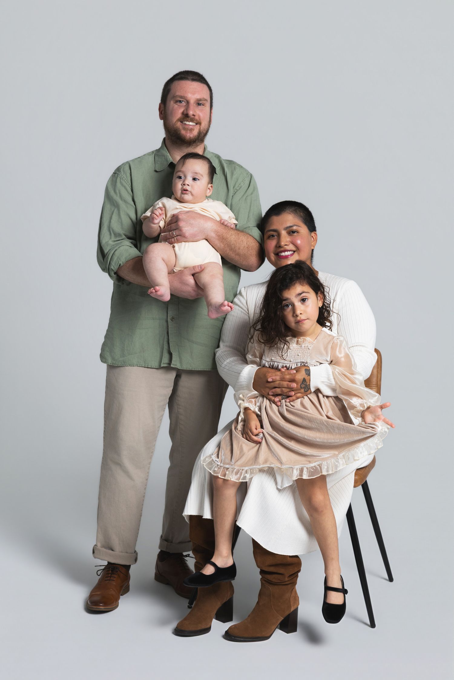 Family portrait with dad holding baby, mom seated with young daughter, studio background.