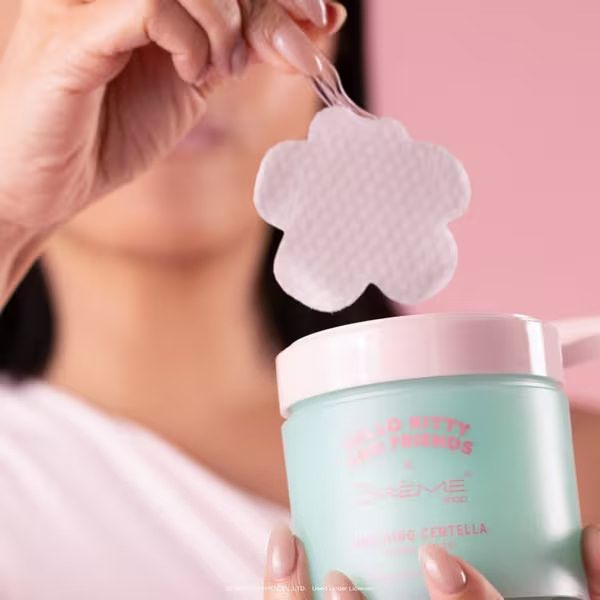 Woman holding a flower-shaped exfoliating pad above a mint green skincare toner pad container on pink background.
