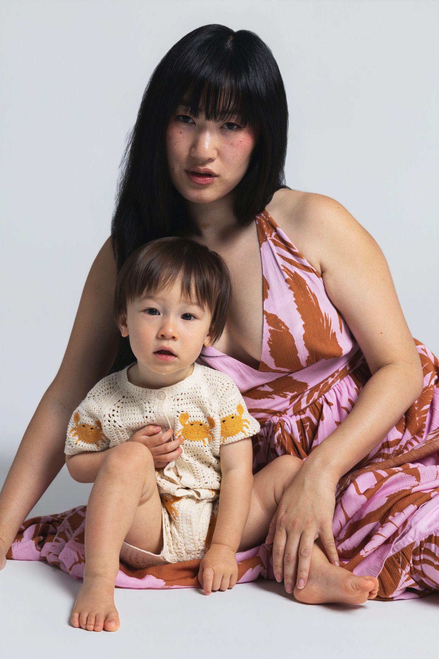 Woman in floral pink dress sits with baby wearing cream knit romper against light gray background.