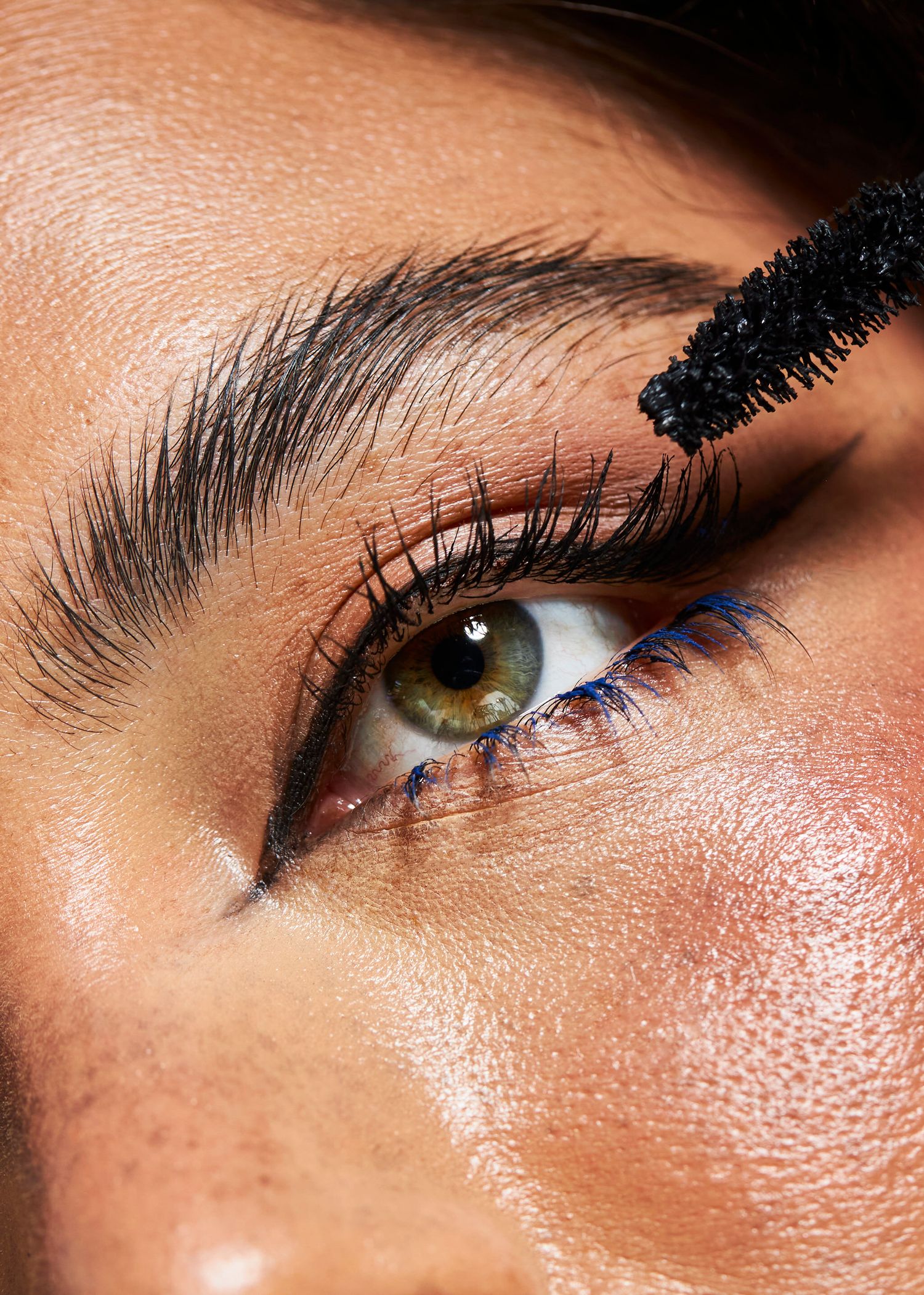 Close-up of a green eye with long lashes being applied with a mascara wand, showcasing bold eye makeup.