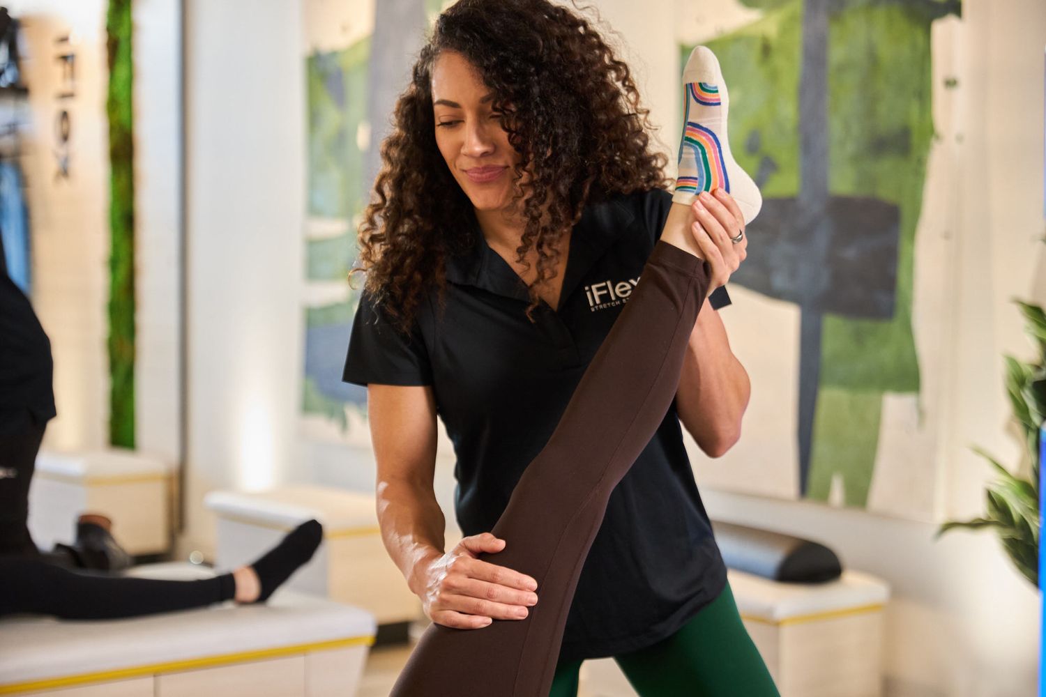 A fitness instructor in a black iFit shirt and green leggings rolls up a brown yoga mat in a bright studio.