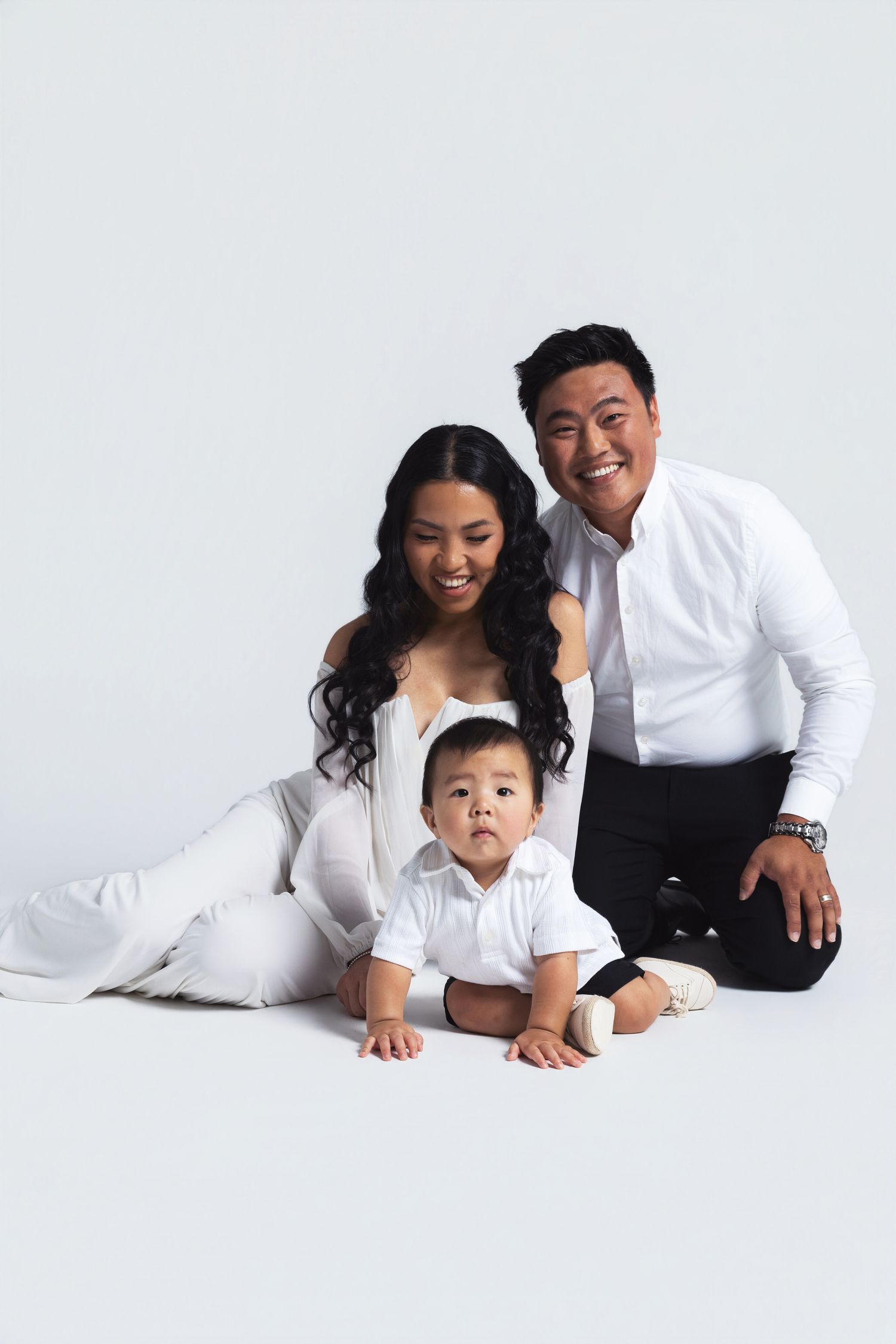 A smiling Asian family in white outfits poses together, with parents sitting behind their cute baby on a white background.
